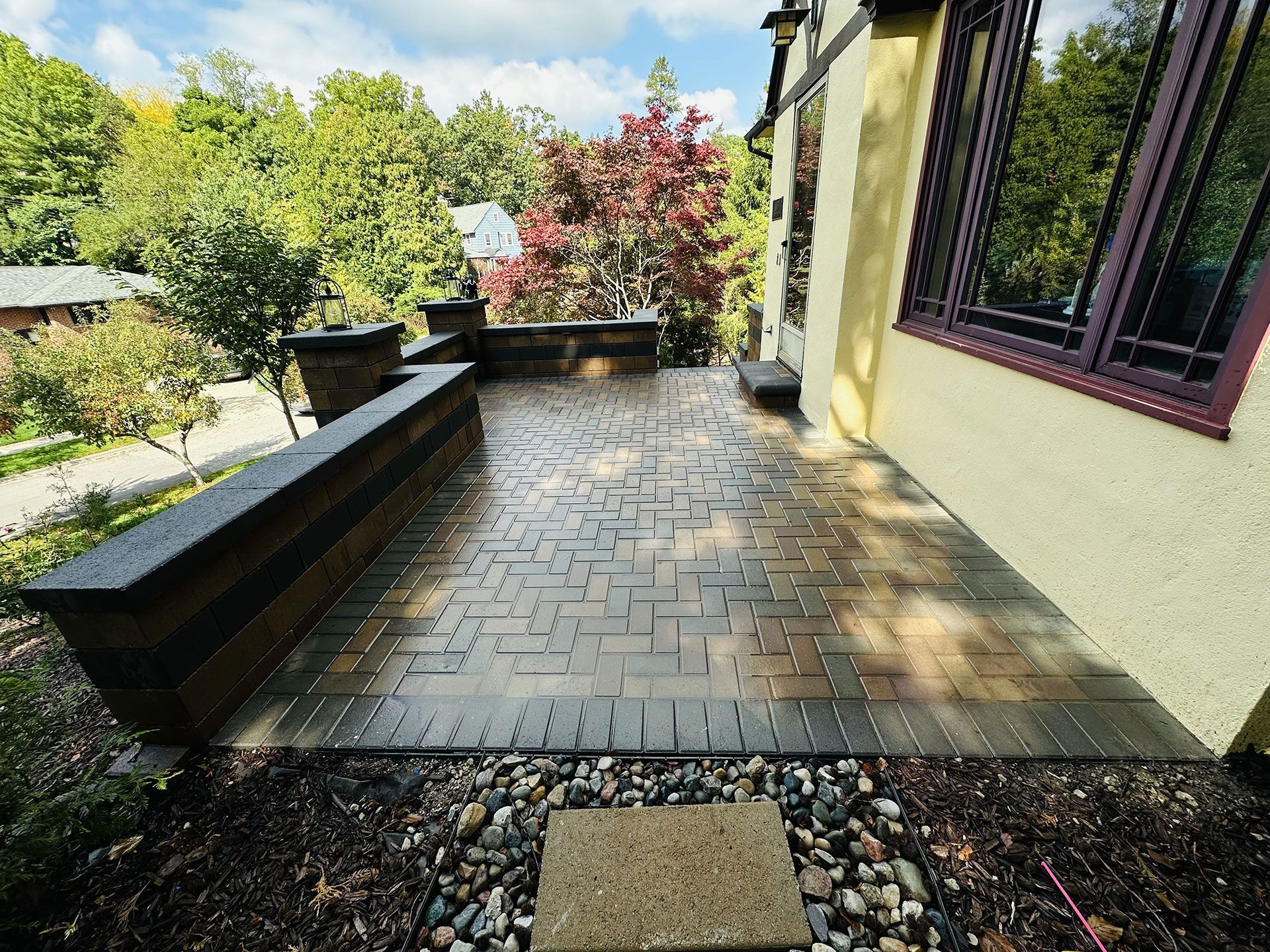 Stone patio outside a building. Brown, rectangular paving stones; wood trim; gravel and landscaping.