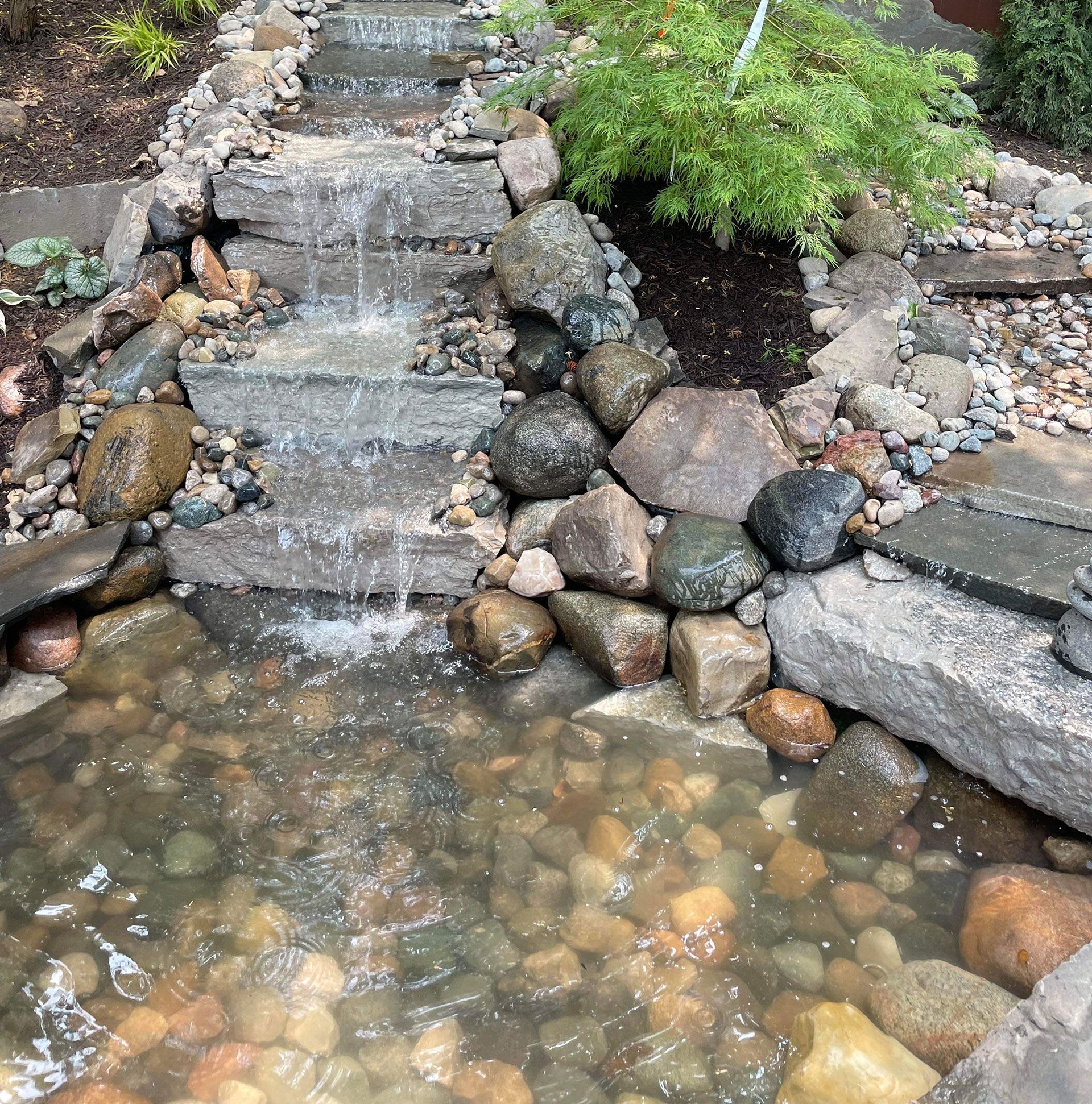 Water cascading down stone steps into a pond filled with rocks; small tree and greenery beside.