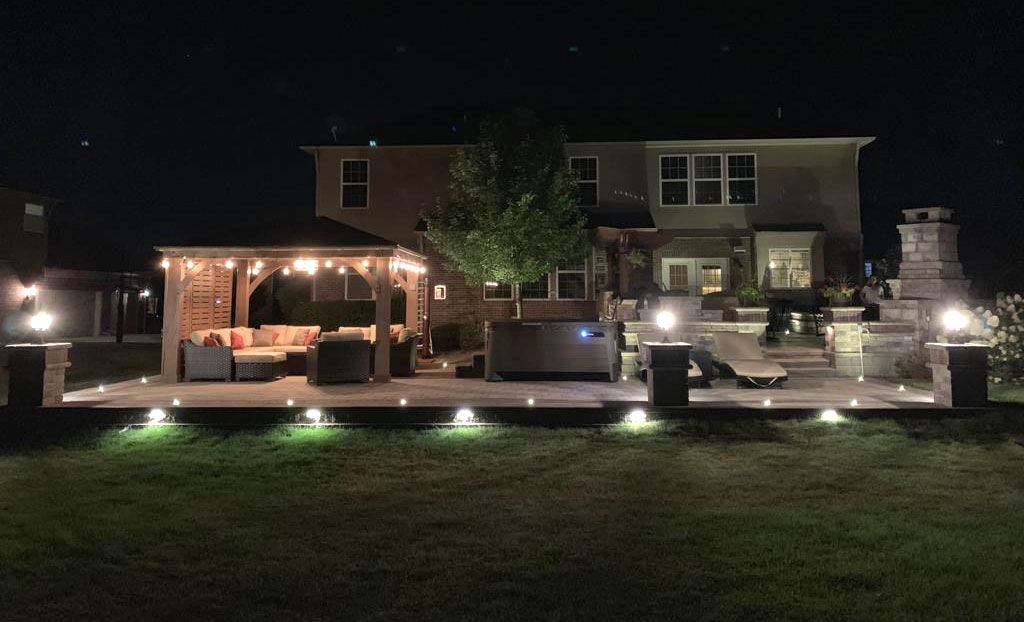 Nighttime backyard patio with gazebo, hot tub, and outdoor lighting, beside a two-story house.