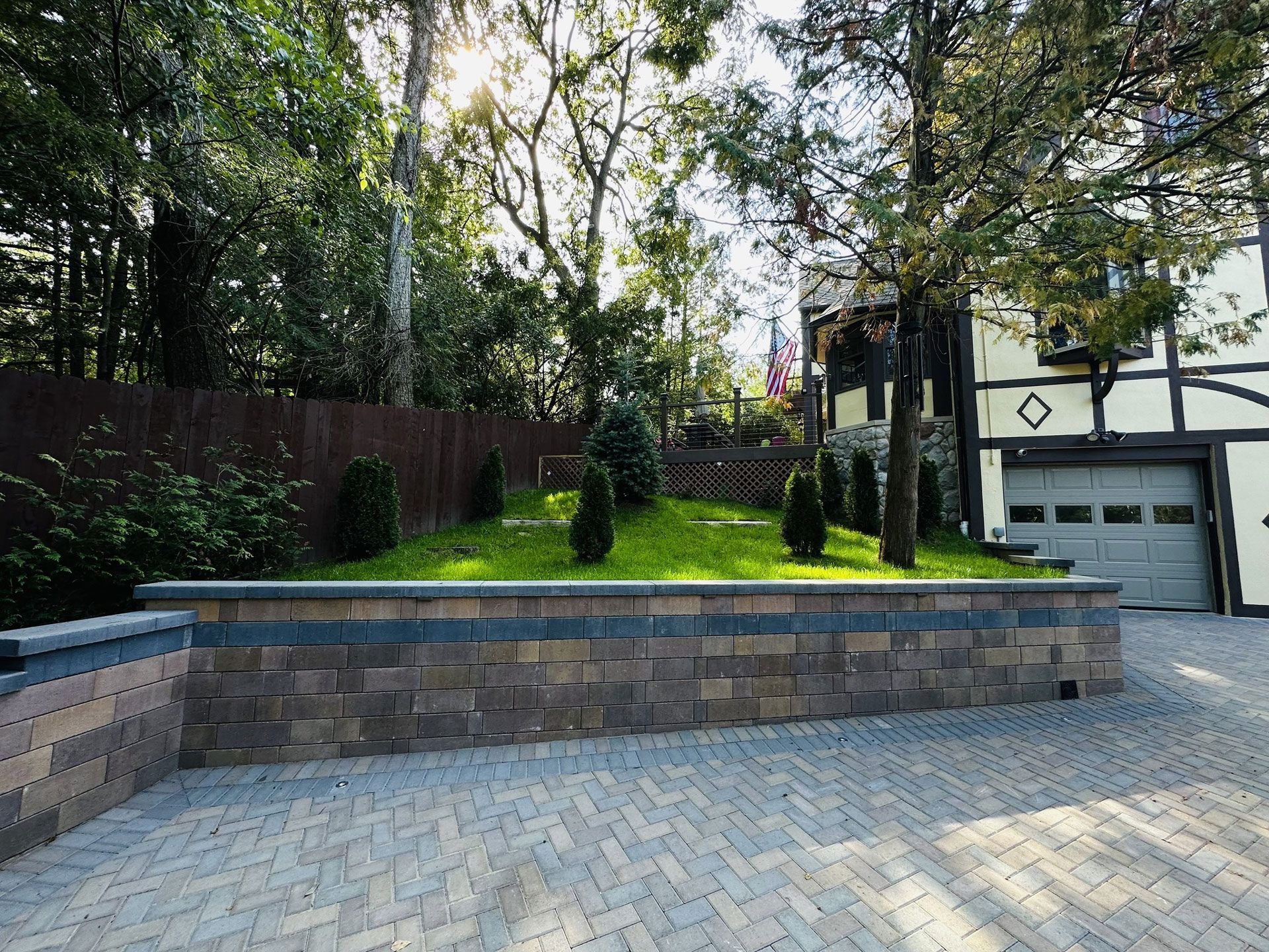 Stone retaining wall with a grassy hill and trees, next to a driveway and Tudor-style house.