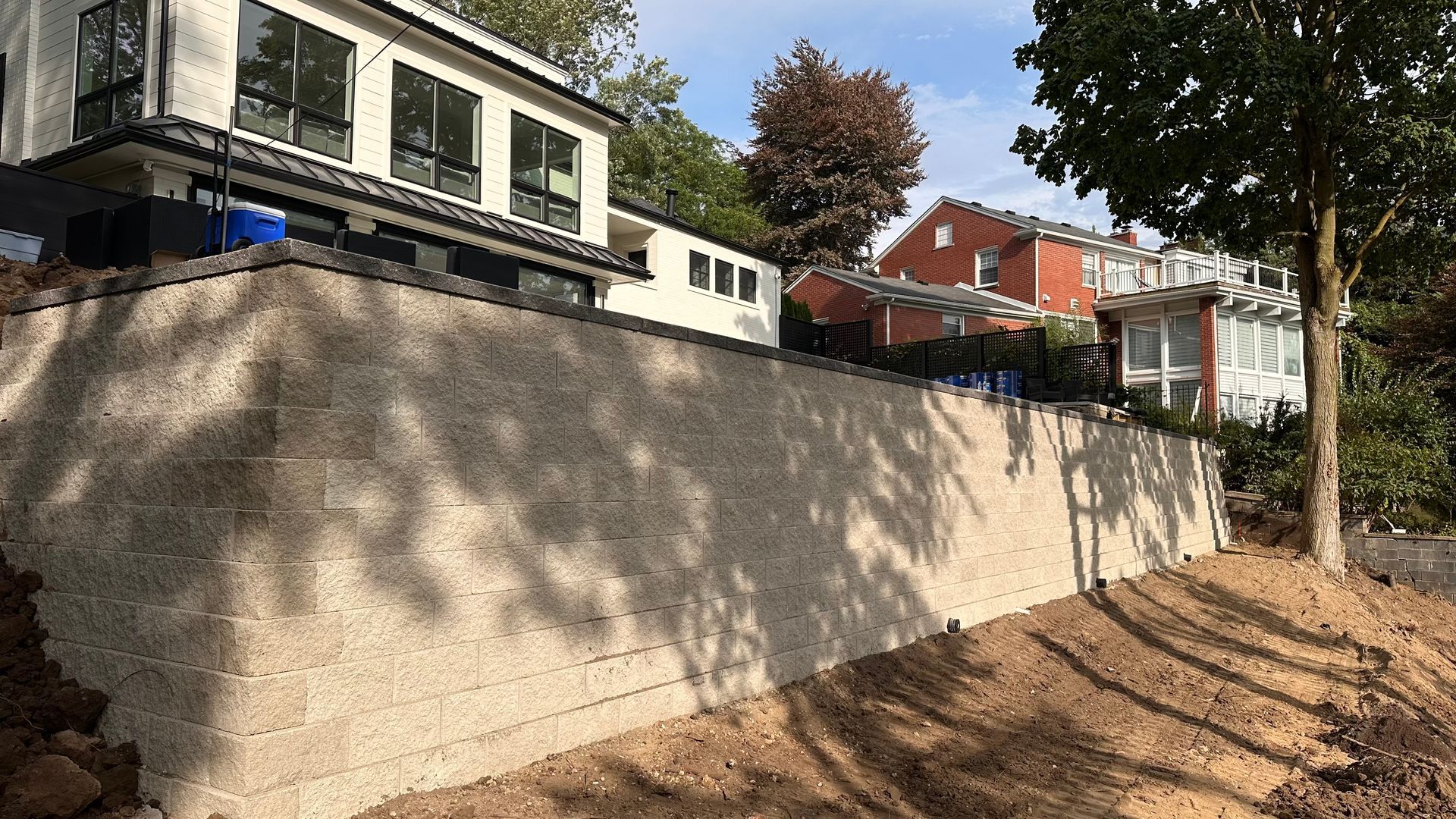 Beige retaining wall in front of a modern house, dirt slope in foreground, with other houses and trees in background.