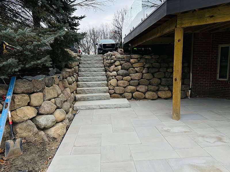 Stone steps lead up to a deck supported by wooden beams, flanked by large rock walls and a paved patio.