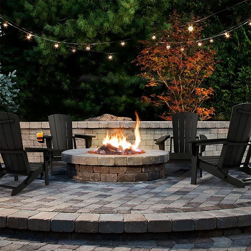 Fire pit with Adirondack chairs on a brick patio, strung with lights, in a backyard.