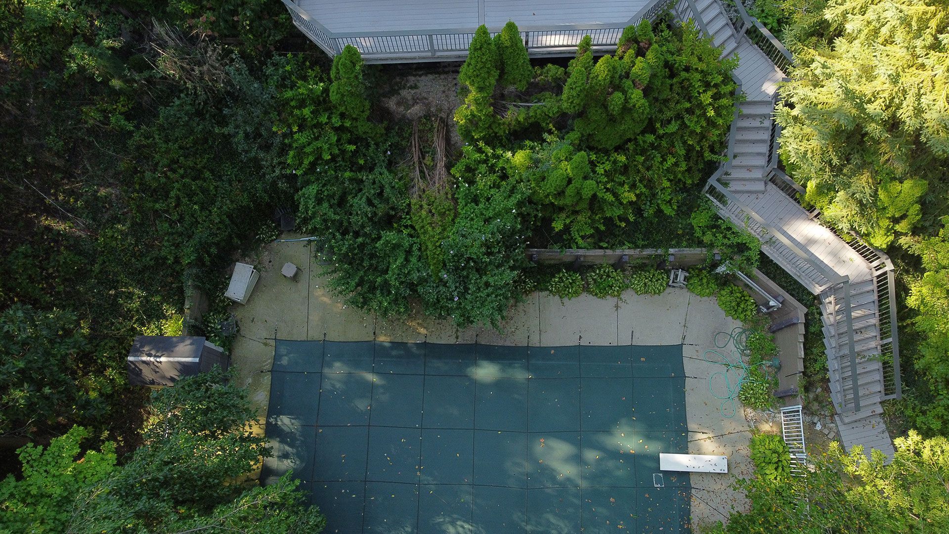 Overhead view of a pool covered with a tarp surrounded by greenery, wooden stairs, and a deck.