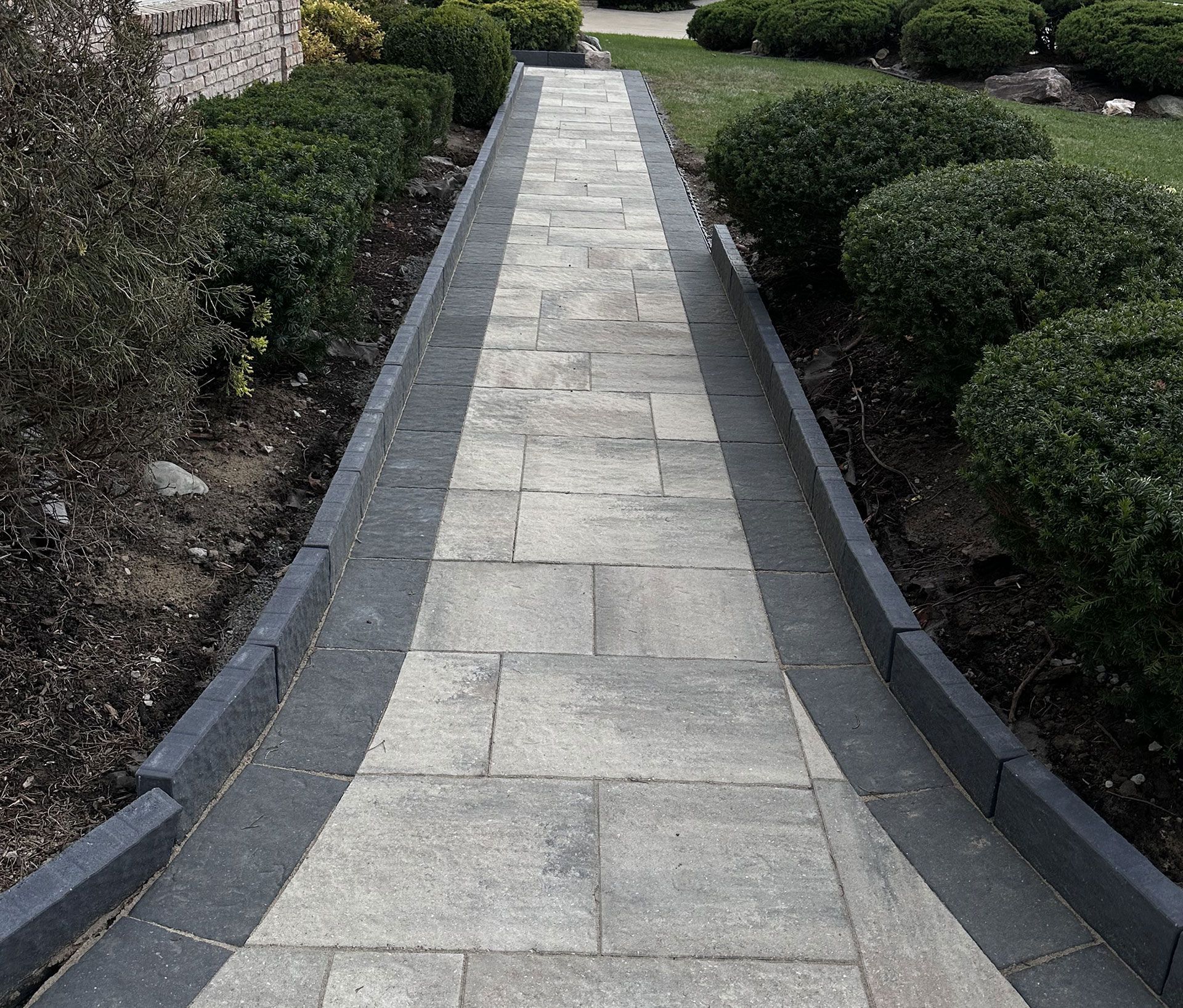 Stone walkway bordered by dark gray curbing and trimmed green bushes.