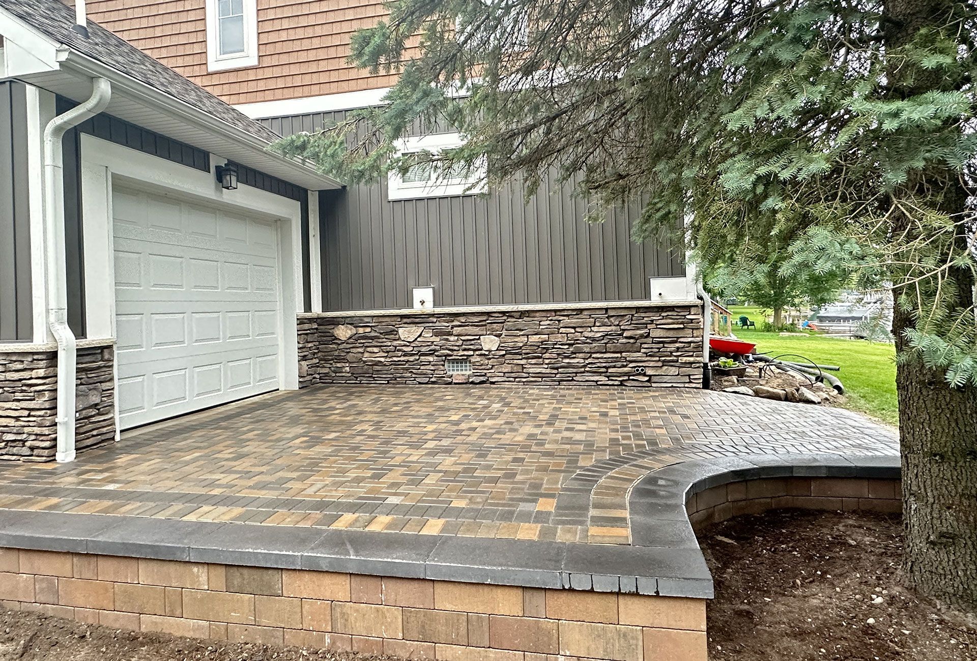 A brick patio with garage, stone wall, and landscaping under a partially cloudy sky.