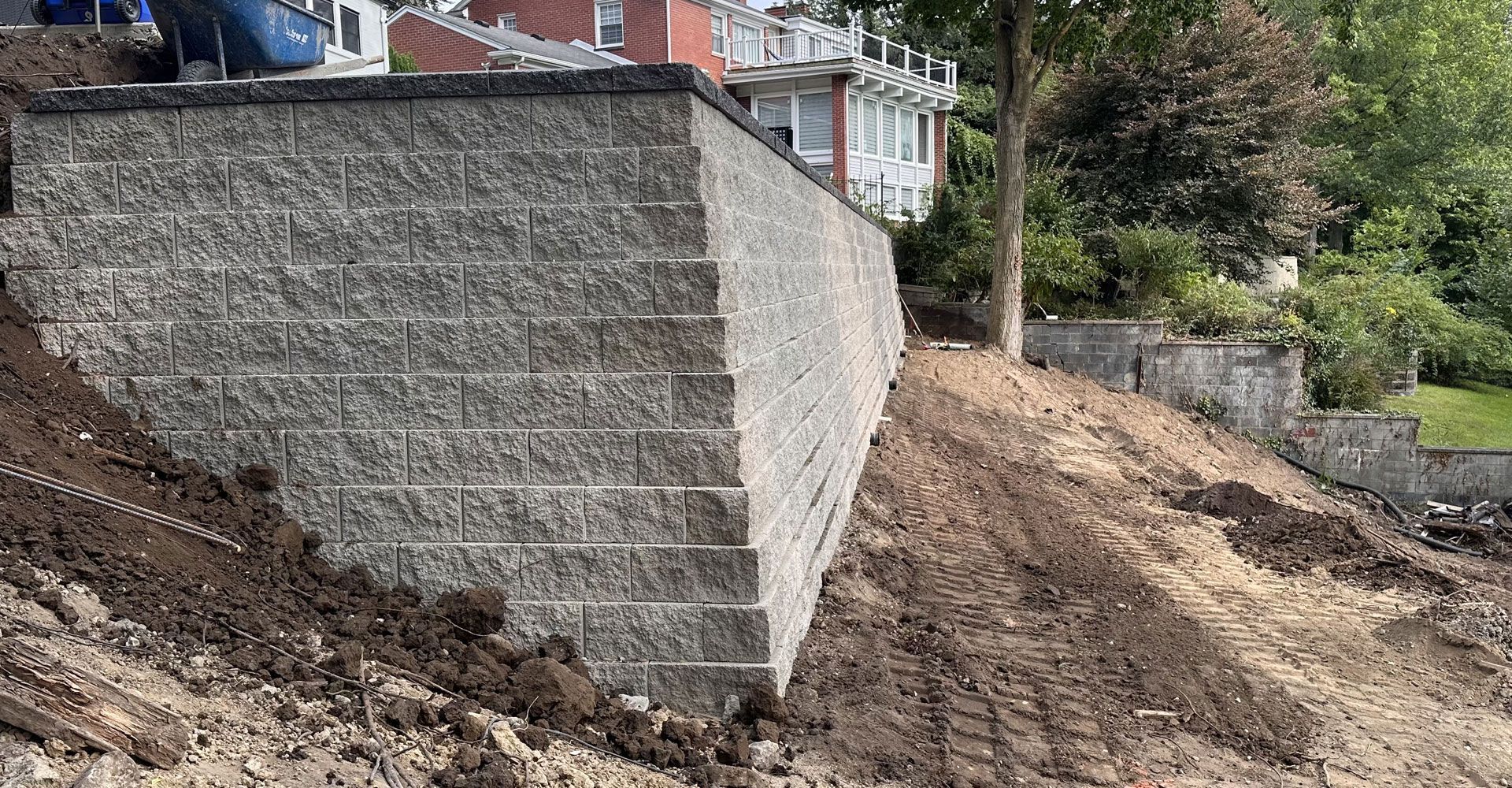 Gray retaining wall built on a hillside. The wall holds back dirt, with a house visible in the background.