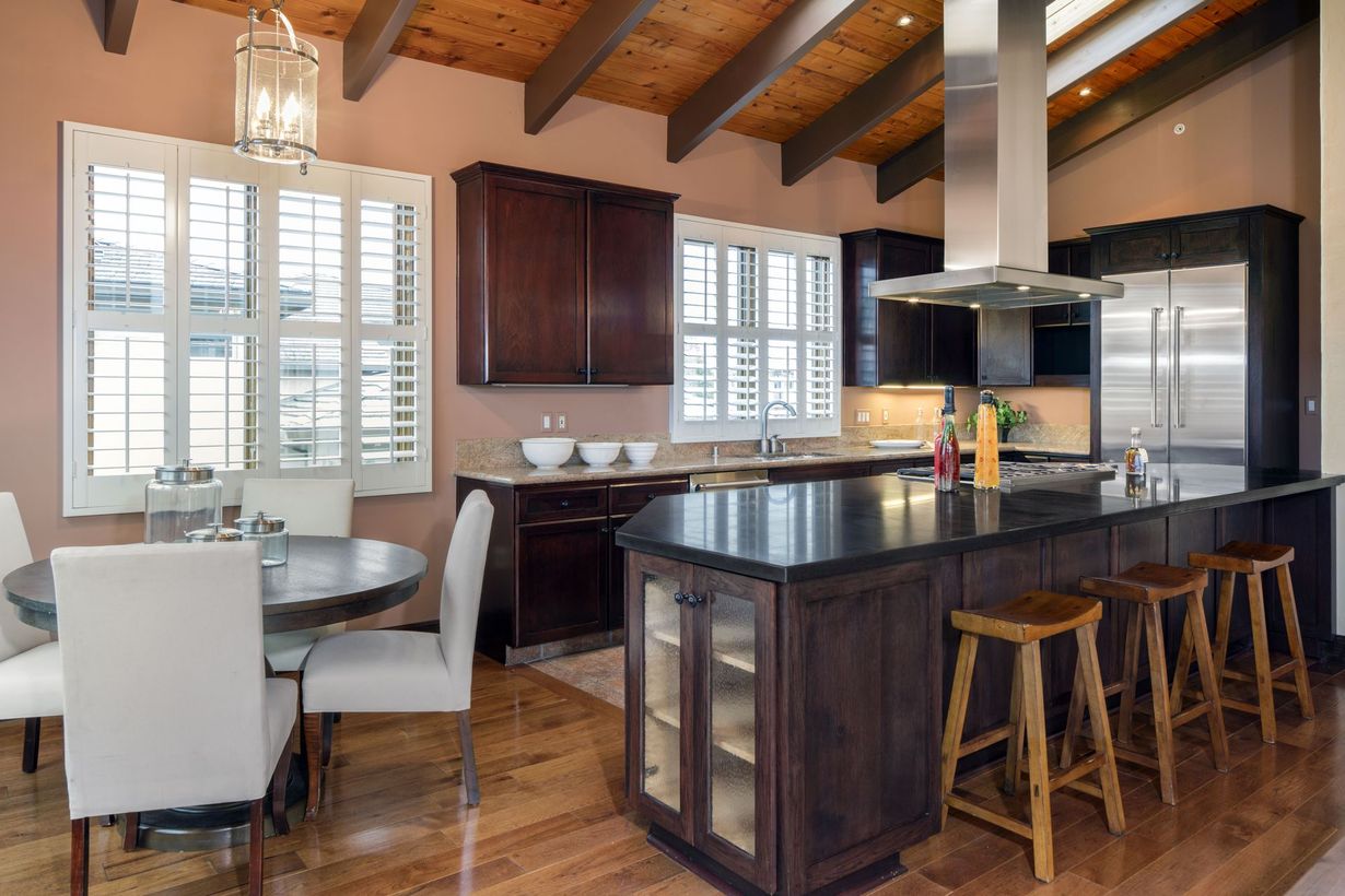 A modern kitchen with dark wood cabinets, a large island, and a dining area. The room has a vaulted ceiling with wooden beams and lots of natural light.