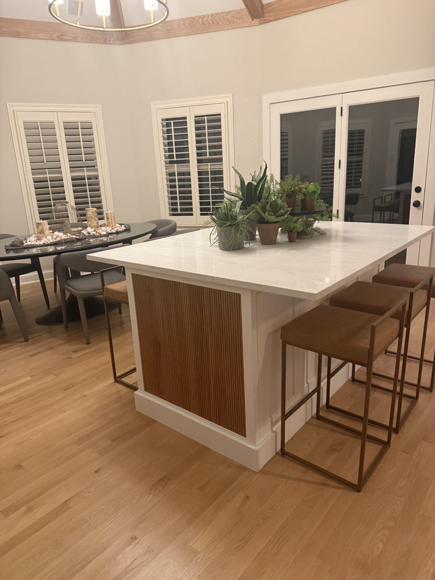Modern kitchen with a white island, wood accents, potted plants, and bar stools on hardwood floors