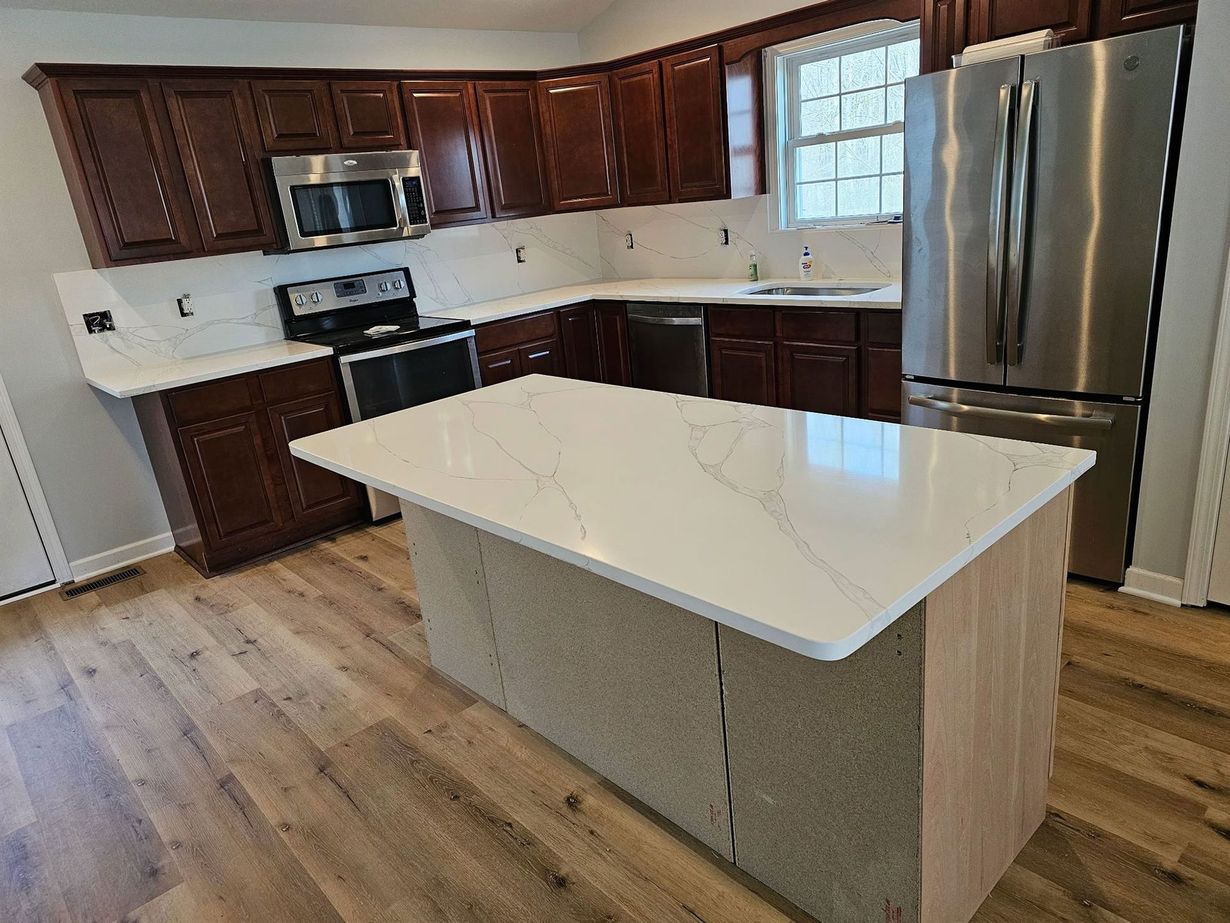 Kitchen with dark wood cabinets, white countertops, and a stainless steel refrigerator. A light wood floor and an island with a white countertop are in the center.