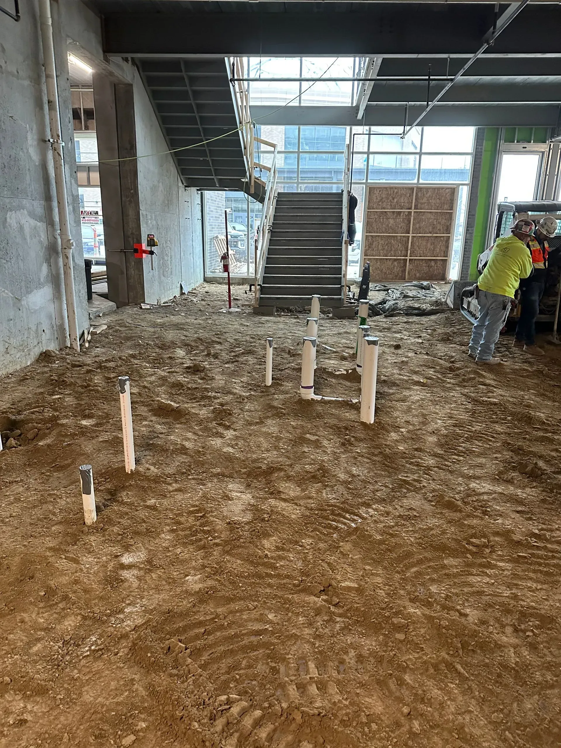 Construction site interior. Plumbing pipes and dirt floor. Staircase and workers visible.