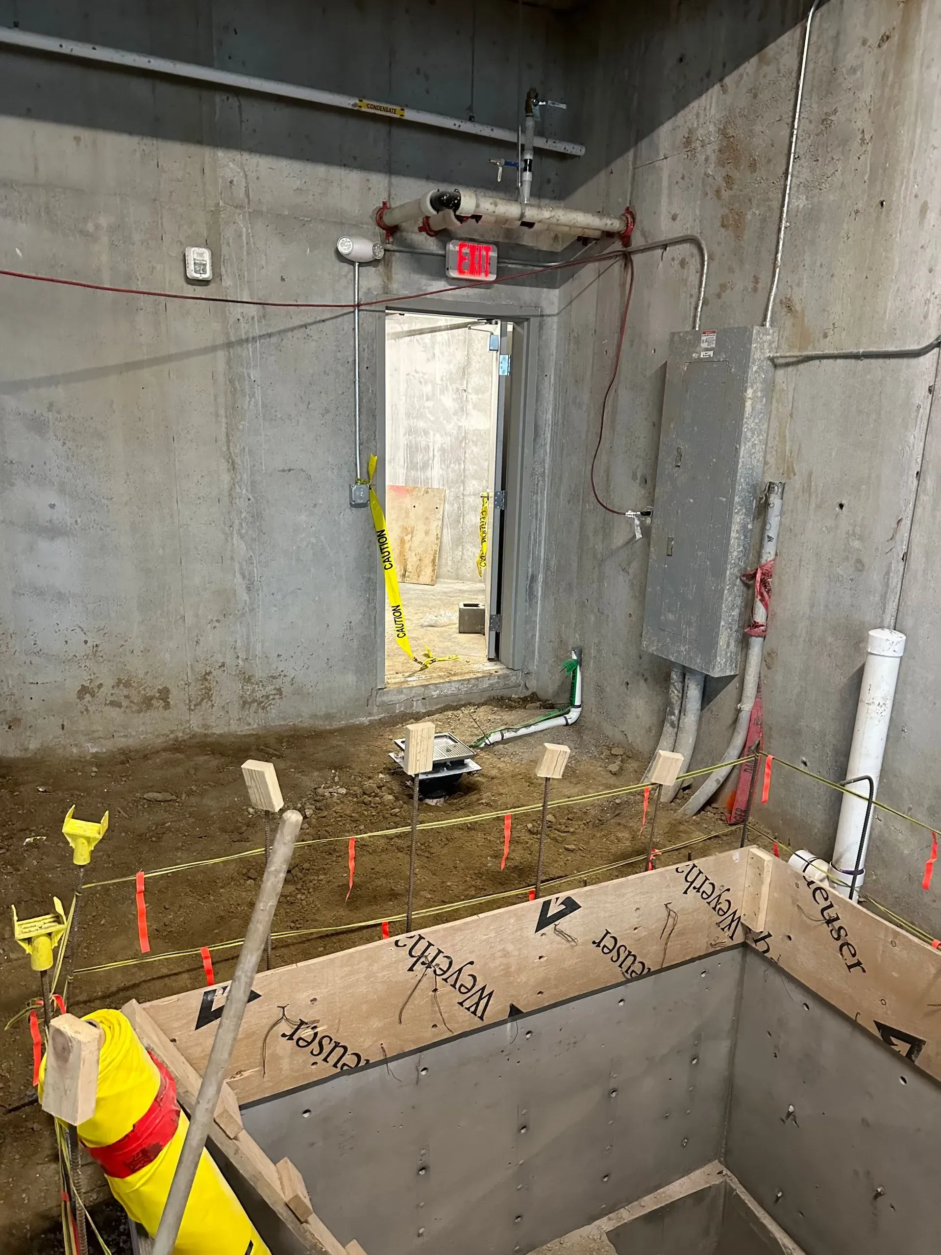 Construction site interior with concrete walls, open doorway, electrical panel, and exposed pipes.