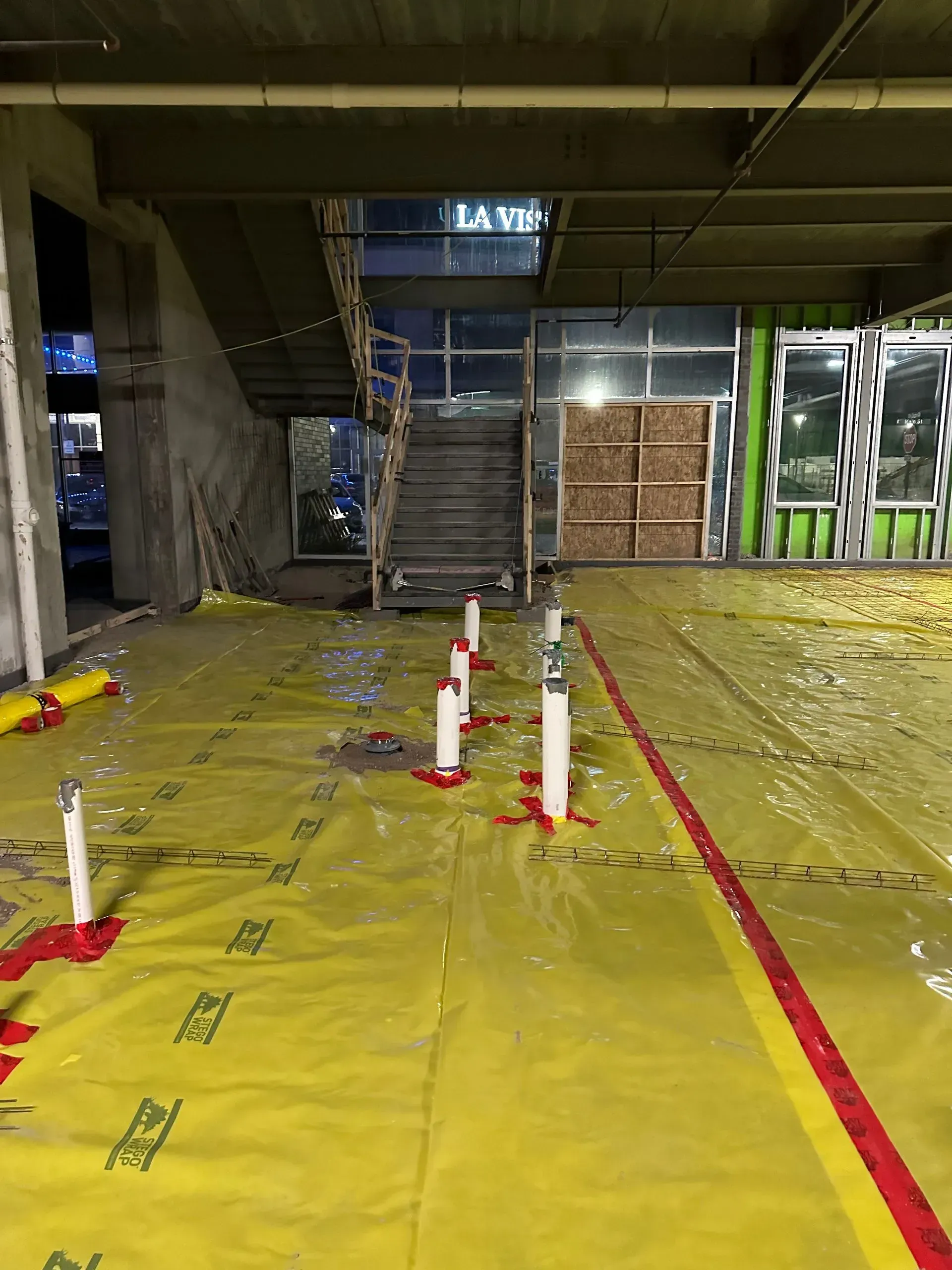 Construction site interior; yellow floor covering with white pipes, stairs in background.