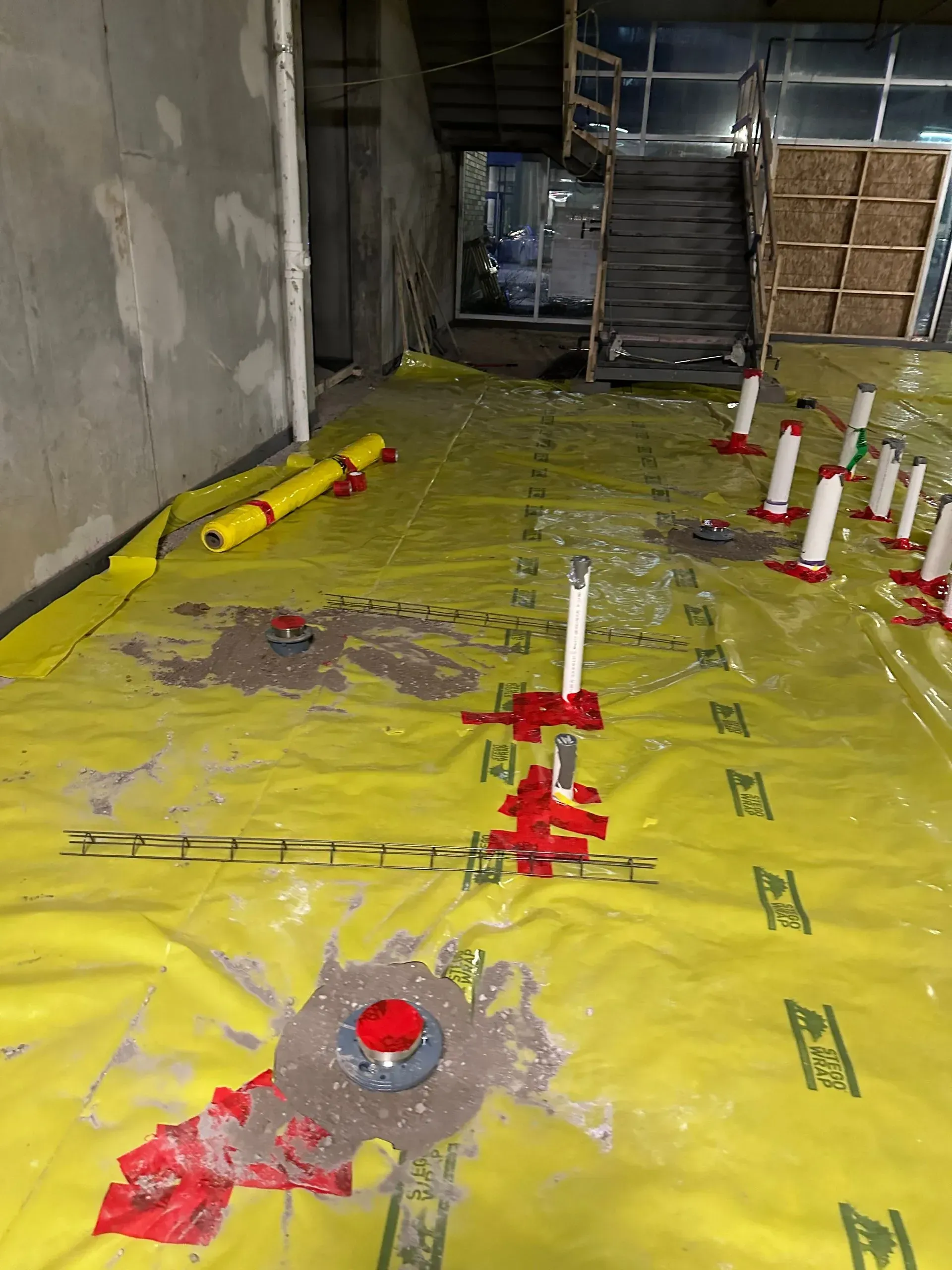 Yellow sheeting on a construction site floor, with pipes, gravel, and chicken feeders. Stairway in background.