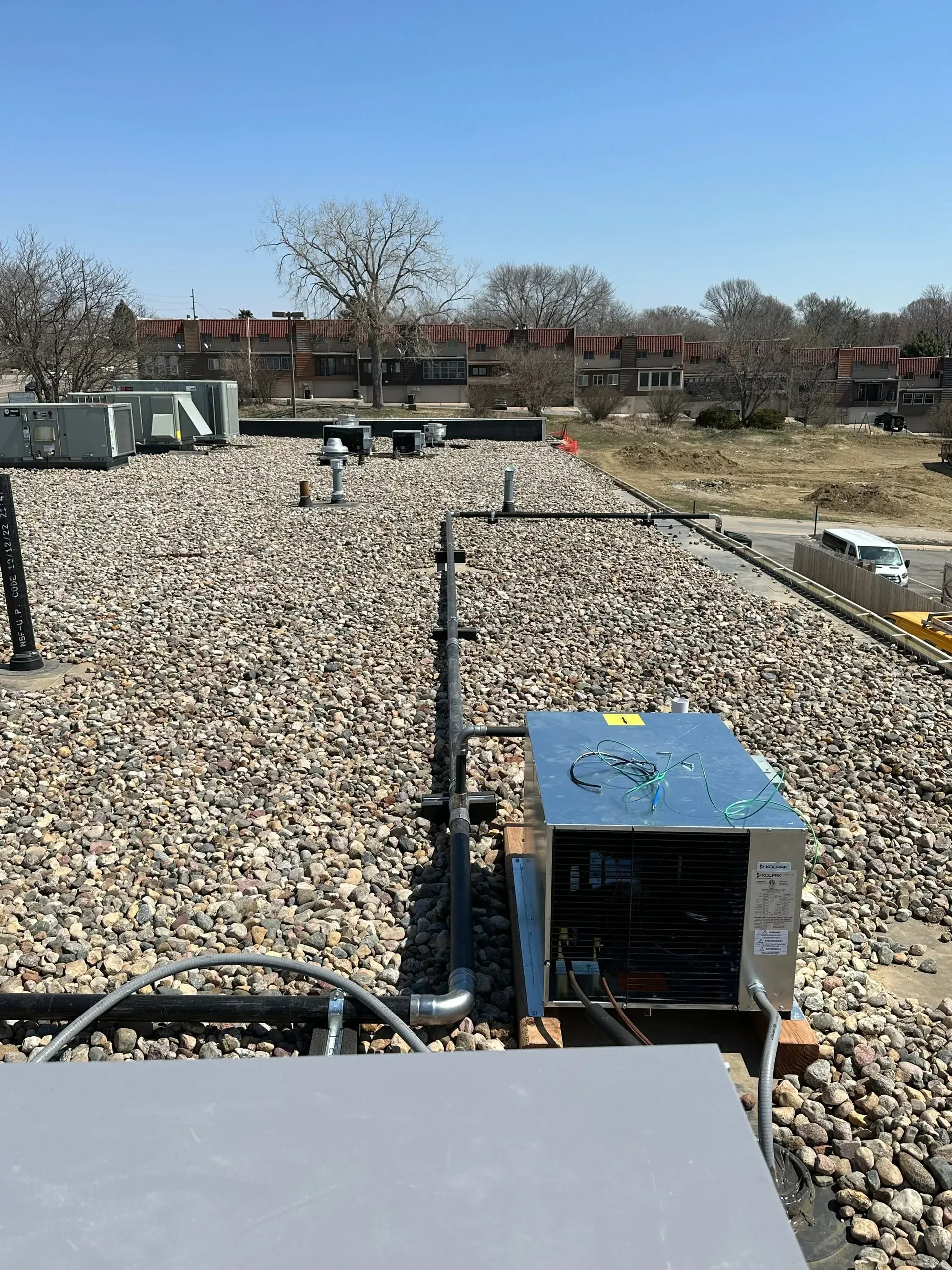 Rooftop with gravel, HVAC units, pipes, and a view of buildings on a clear, sunny day.