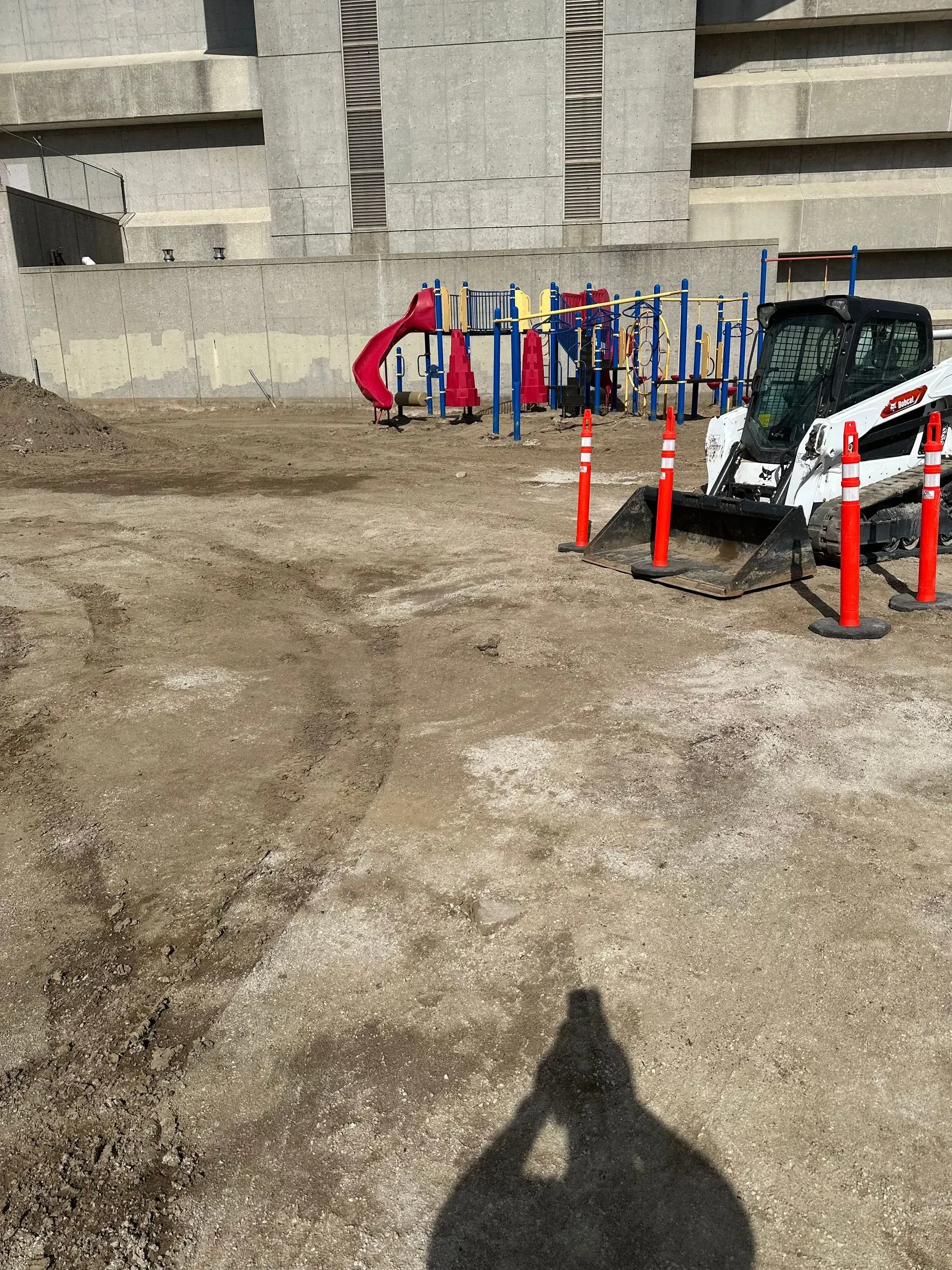 Playground under construction. Bobcat tractor, orange cones, and playground equipment in front of a concrete wall.