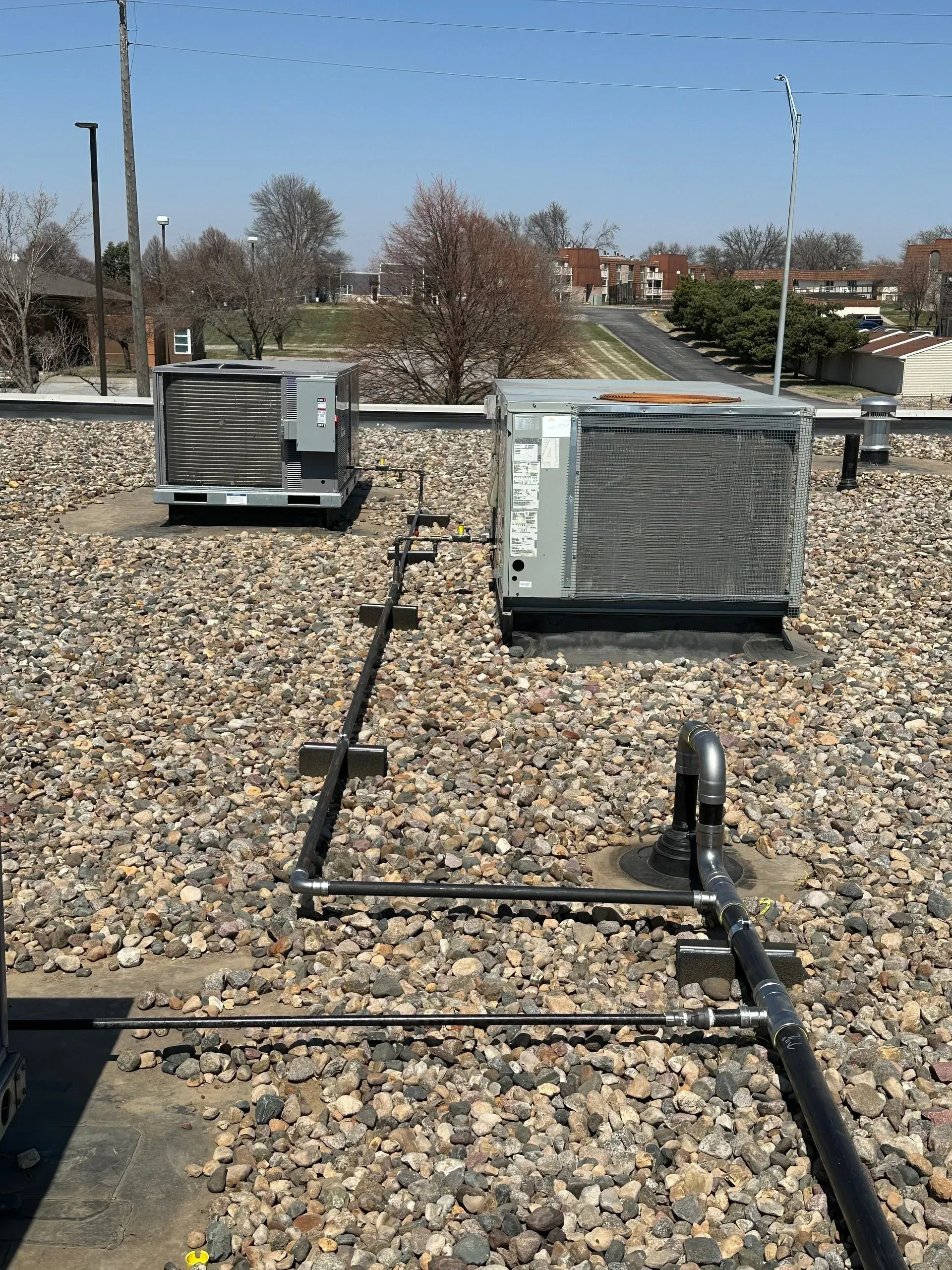 Rooftop scene with two air conditioning units, pipes, and gravel surface under a blue sky.