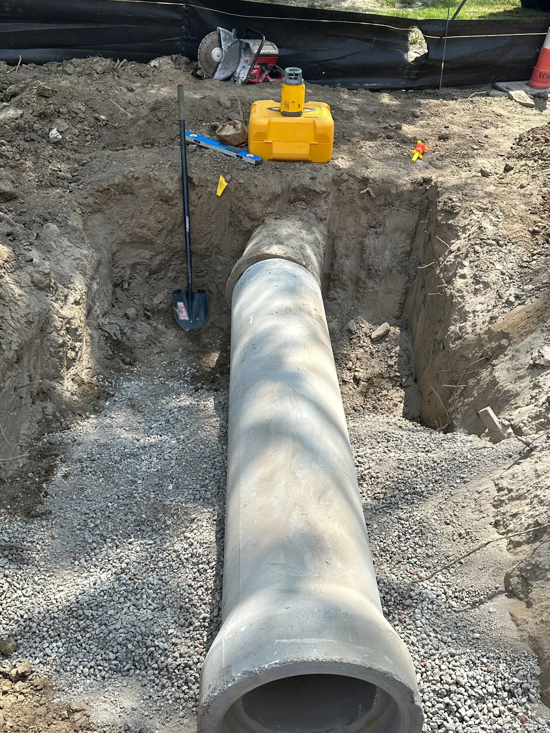 A concrete pipe in a trench with construction tools and gravel.