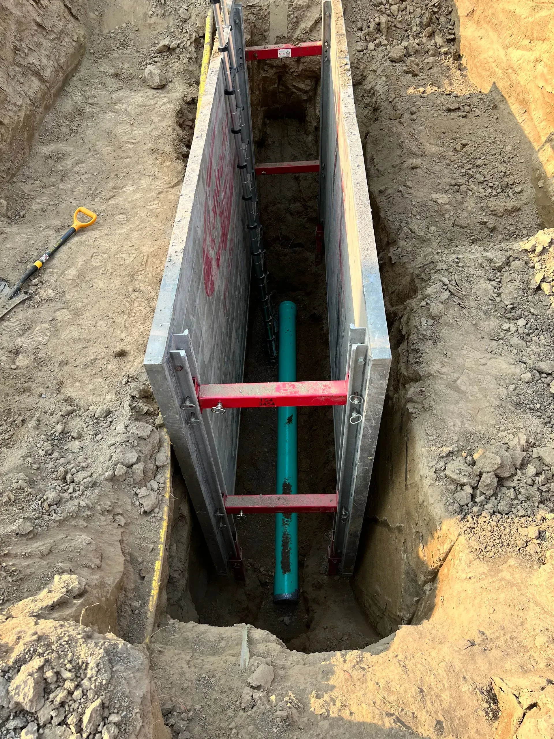 Trench with aluminum shoring and a green pipe running through it, dug into dirt.
