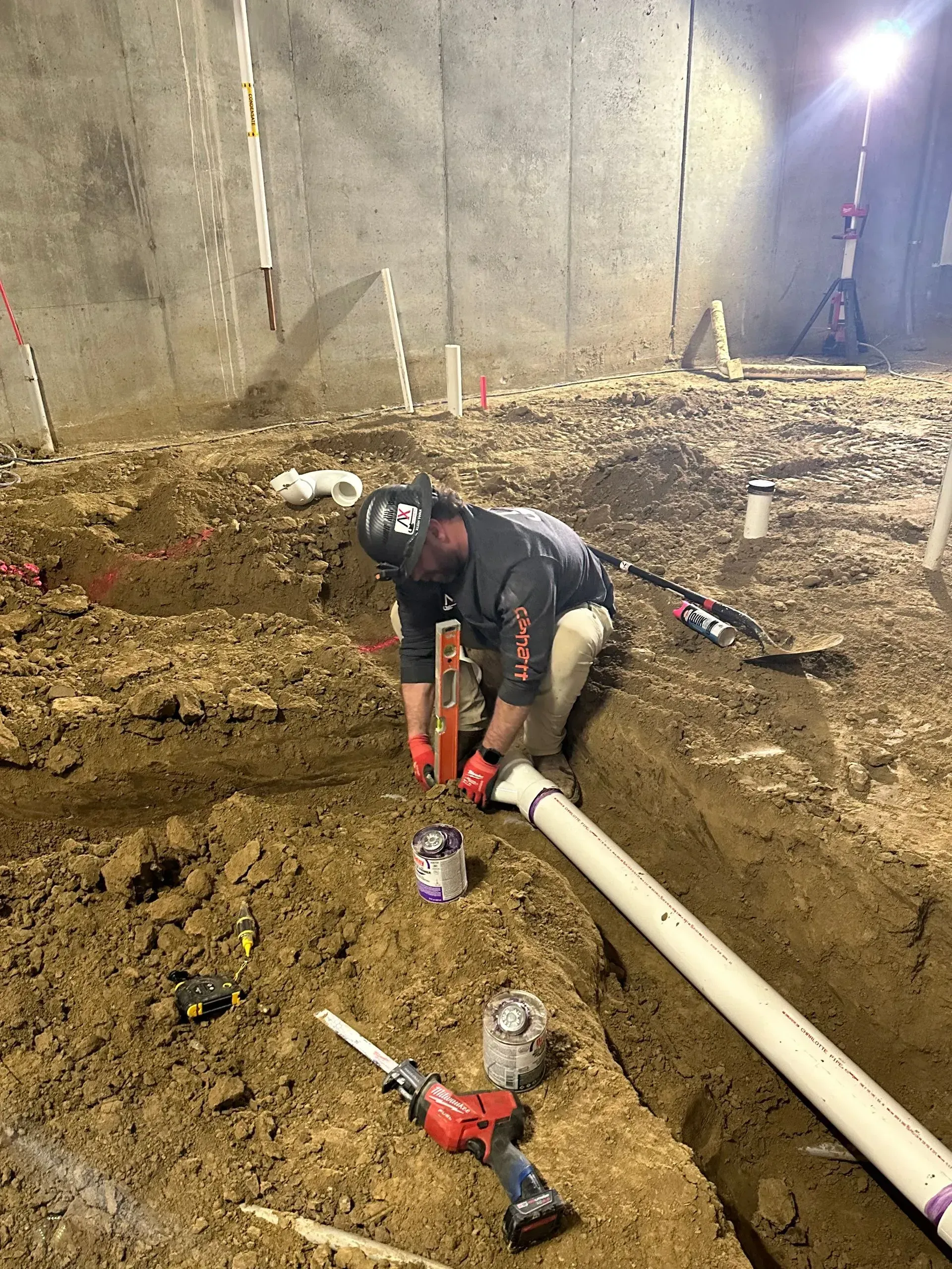 Construction worker using a level to install white pipe in a trench. Concrete wall in background.