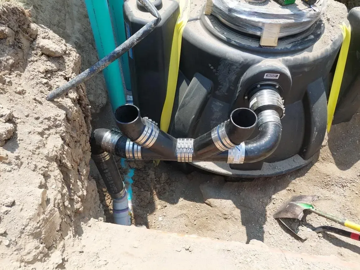 Black septic tank with connecting pipes in a sandy trench. A shovel rests nearby.