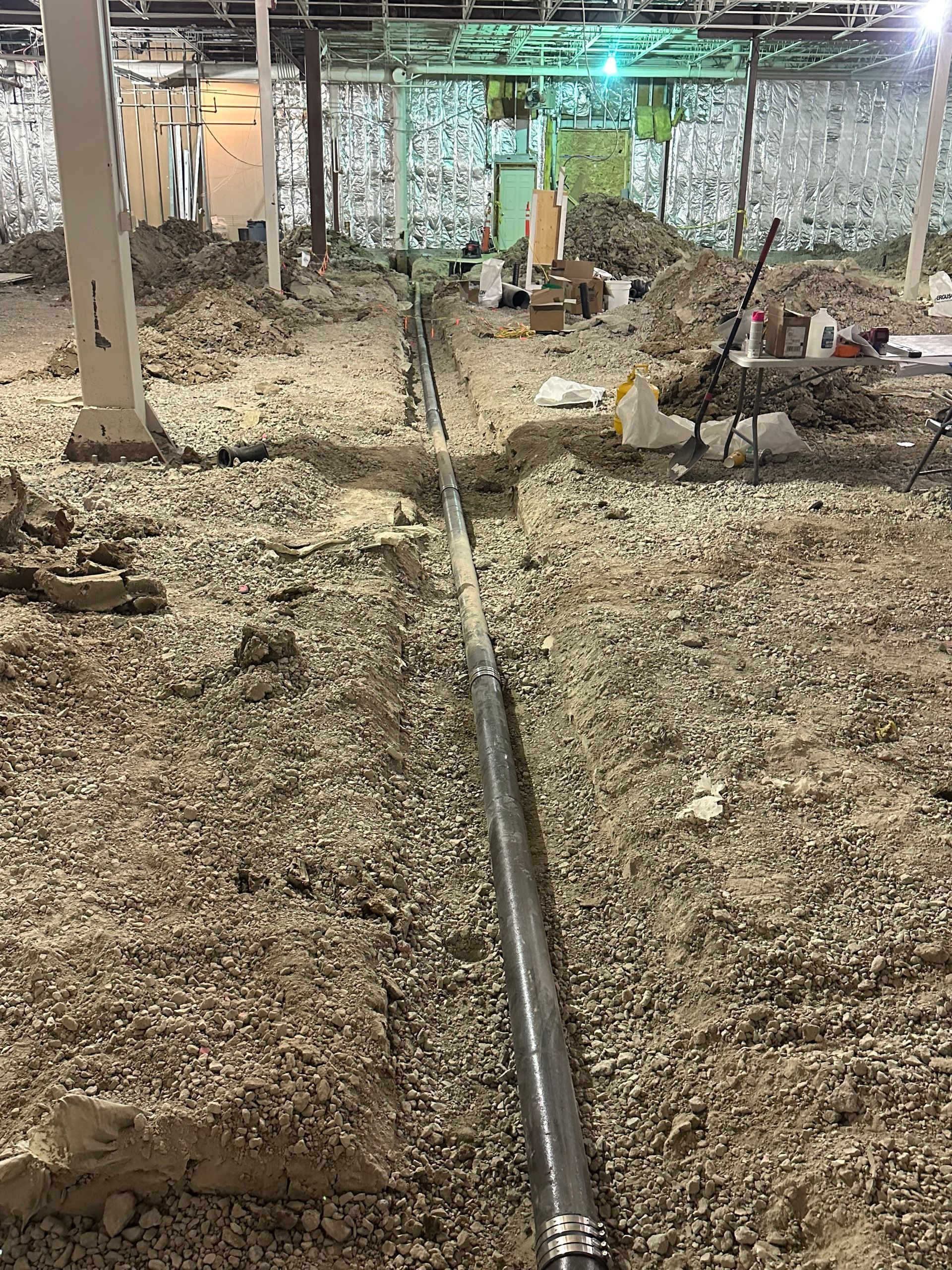 Trench dug with a pipe in the center, surrounded by gravel, inside a building under construction.