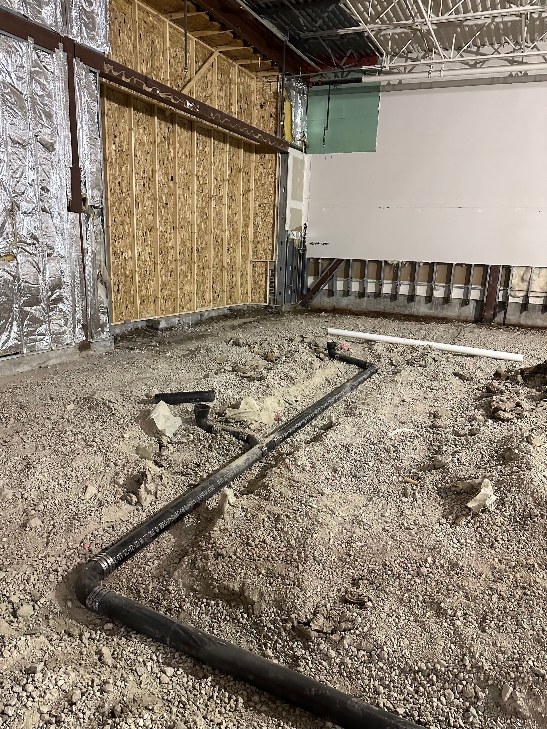 Construction site interior; plumbing pipes on gravel floor, wooden studs and partially finished walls.