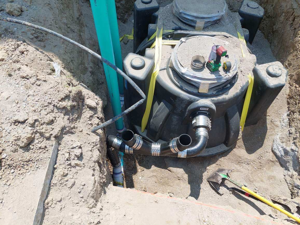 Black septic tank with piping in a dirt trench, surrounded by sand, with a green pipe visible.