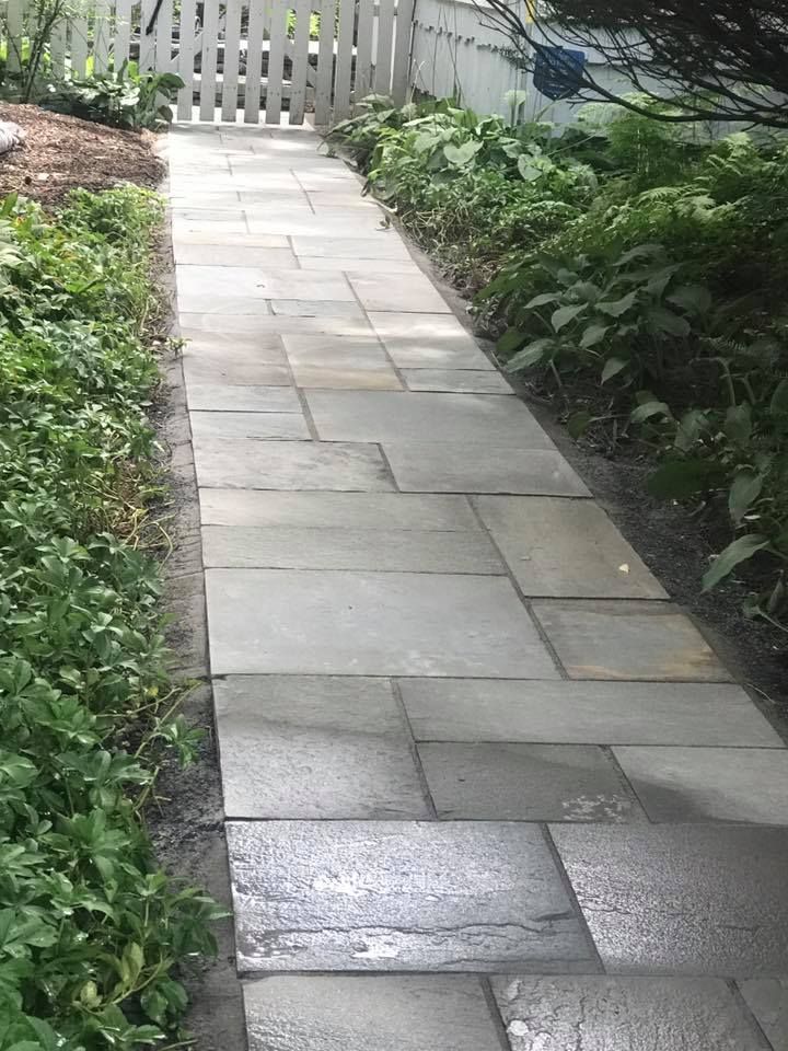 A stone walkway leading to a white picket fence surrounded by plants.