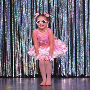 A little girl in a pink tutu and sunglasses is standing on a stage.
