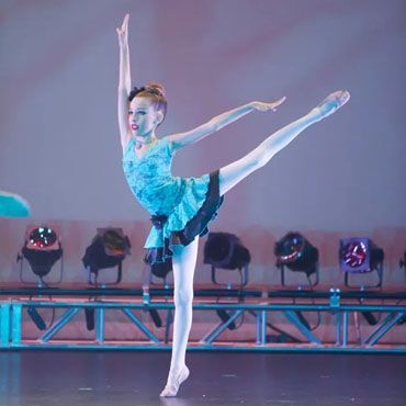 A young ballerina is standing on one leg on a stage.