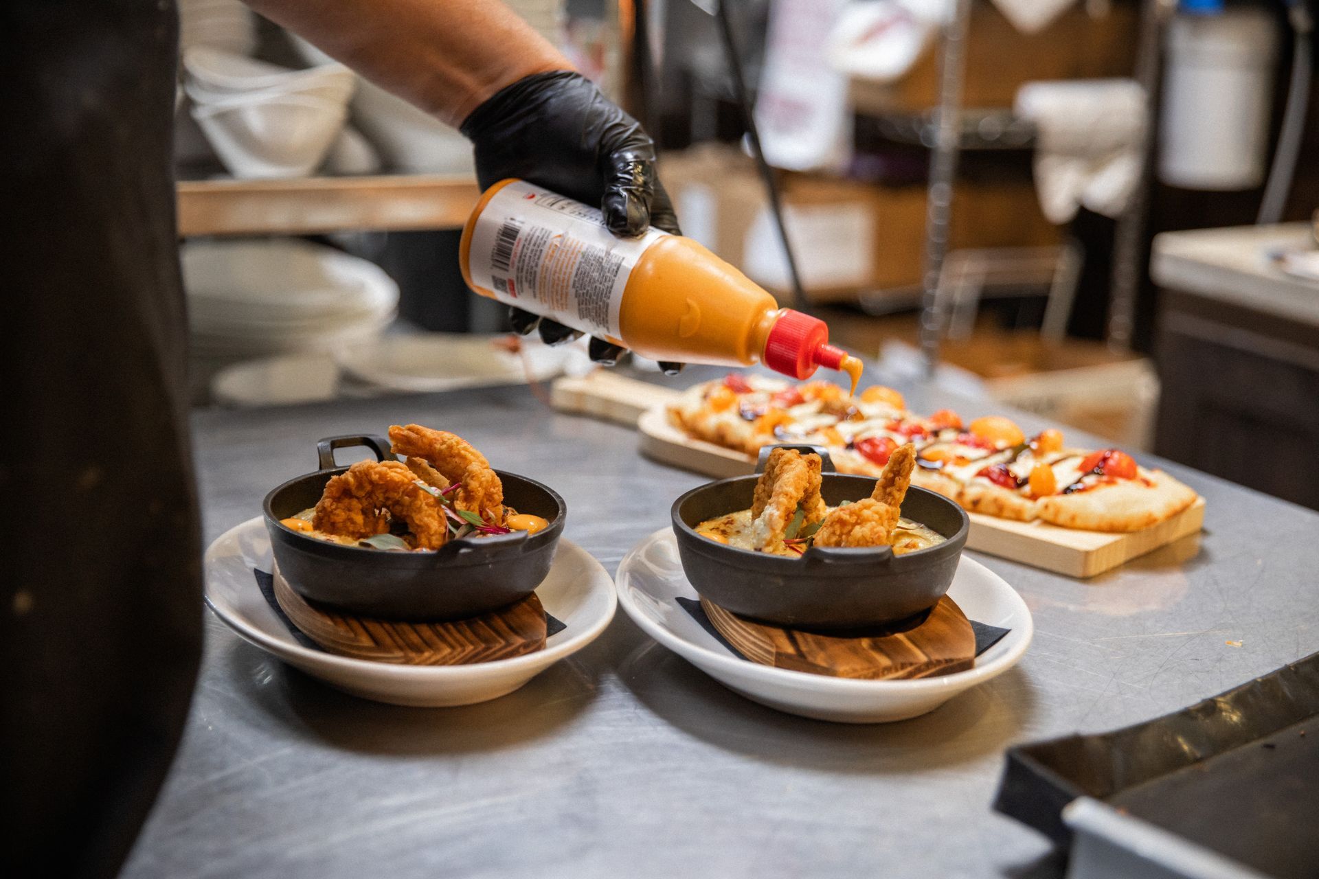 A cook in black gloves pours sauce on two bowls of fried food and a pizza on a metal counter.
