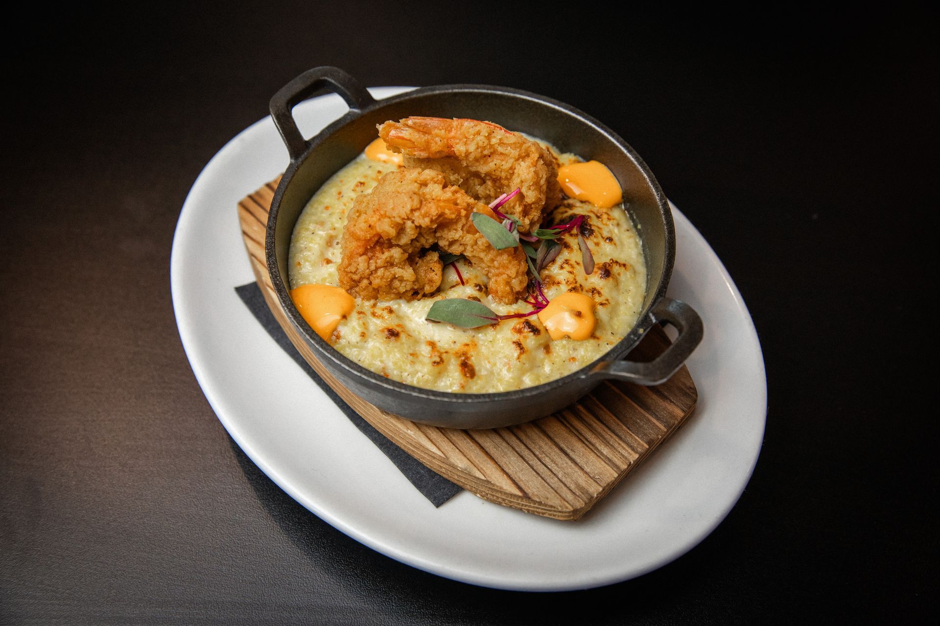 Fried chicken atop creamy grits in a black cast-iron dish, served on a white plate.