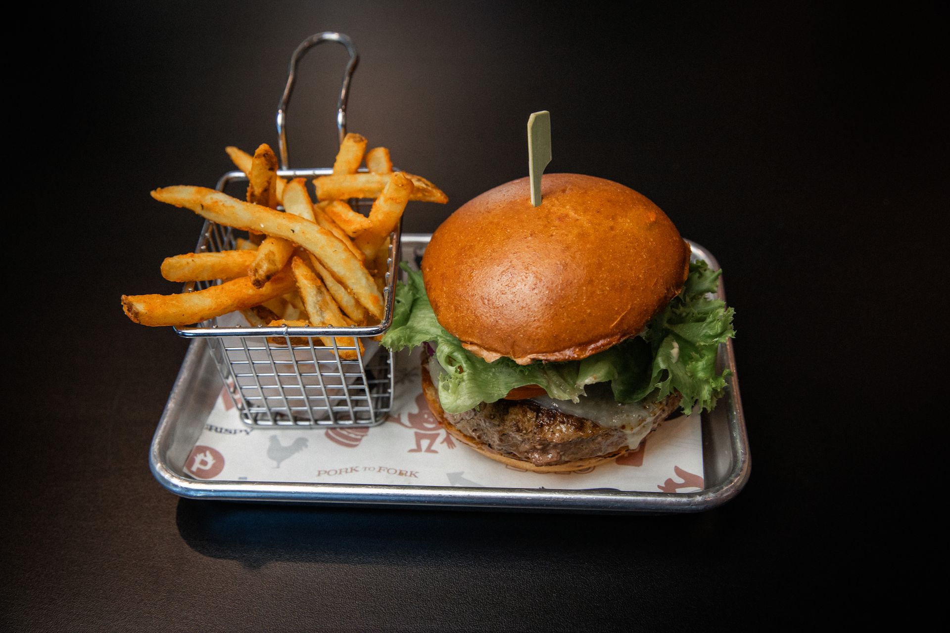 Burger and fries on a tray with a black background. The burger is topped with lettuce and held with a toothpick.