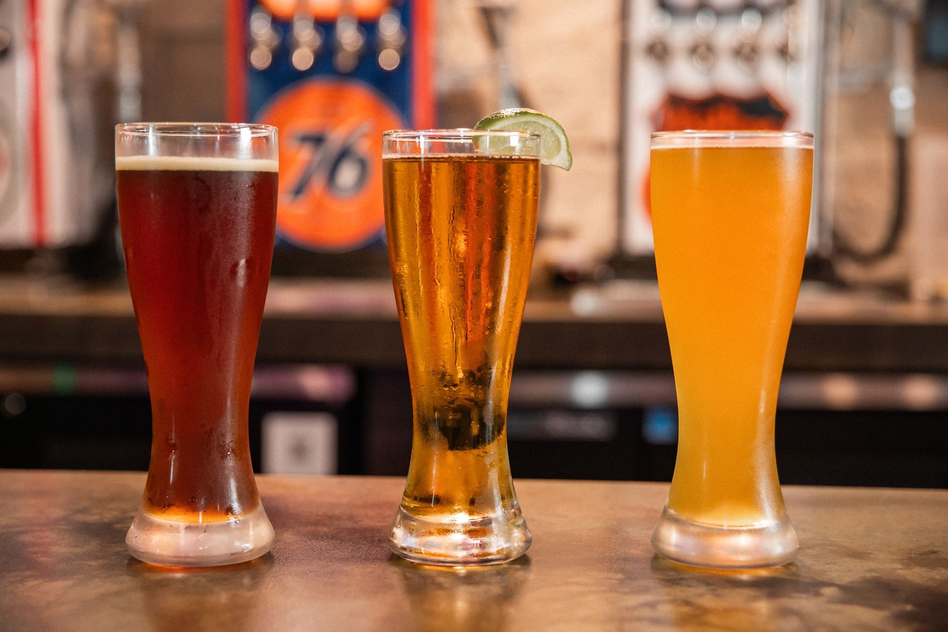 Three glasses of beer on a bar. From left to right: dark brown, light amber with lime, and golden.