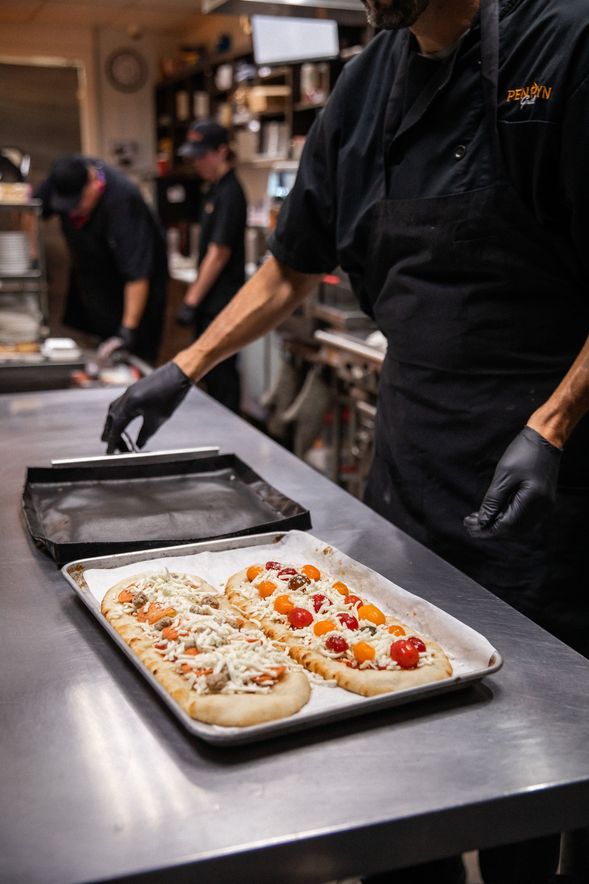 Chef placing flatbreads with toppings on a metal tray in a kitchen. Other workers in background.