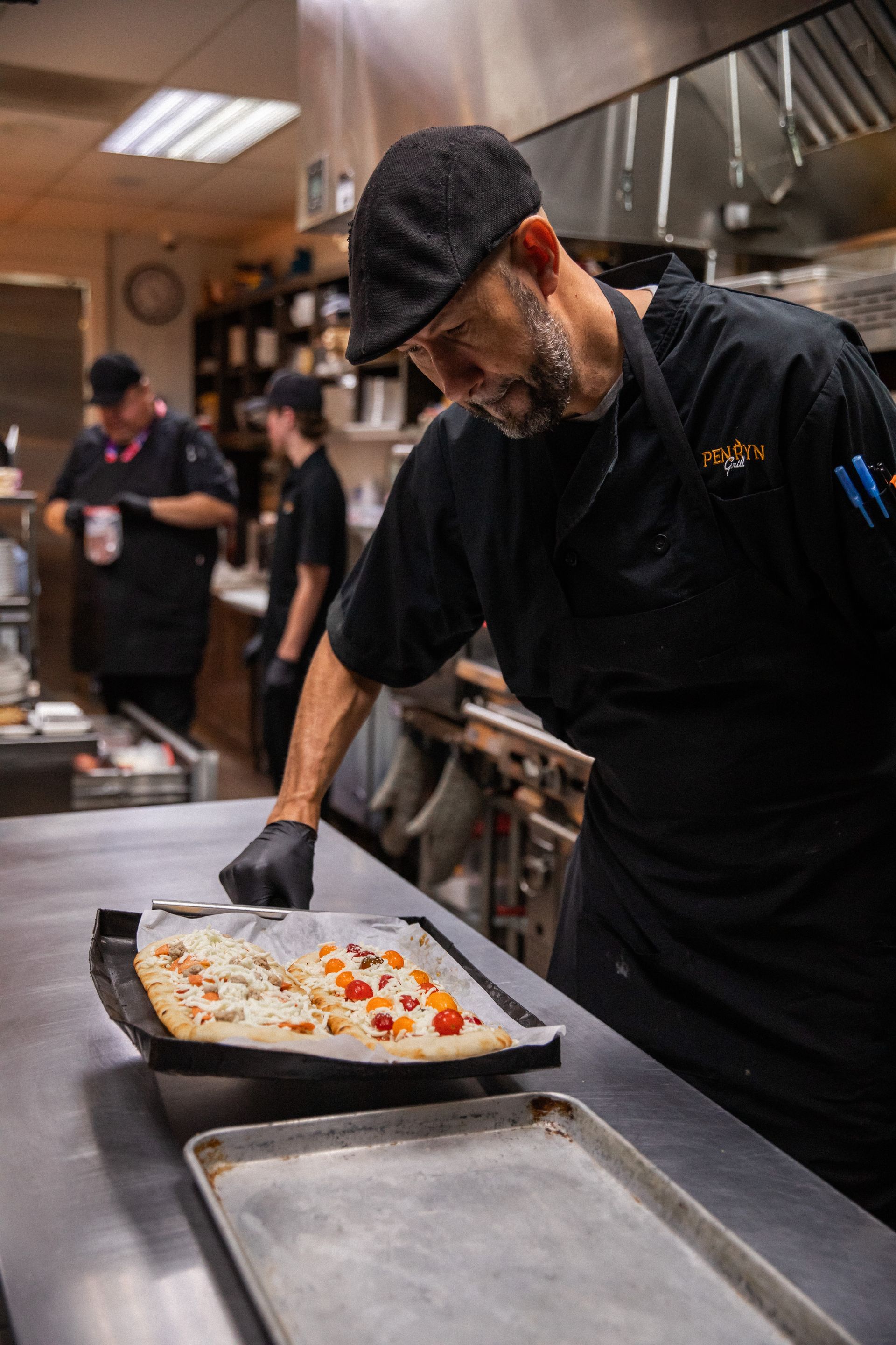 Chef in black uniform examines a flatbread pizza in a restaurant kitchen.