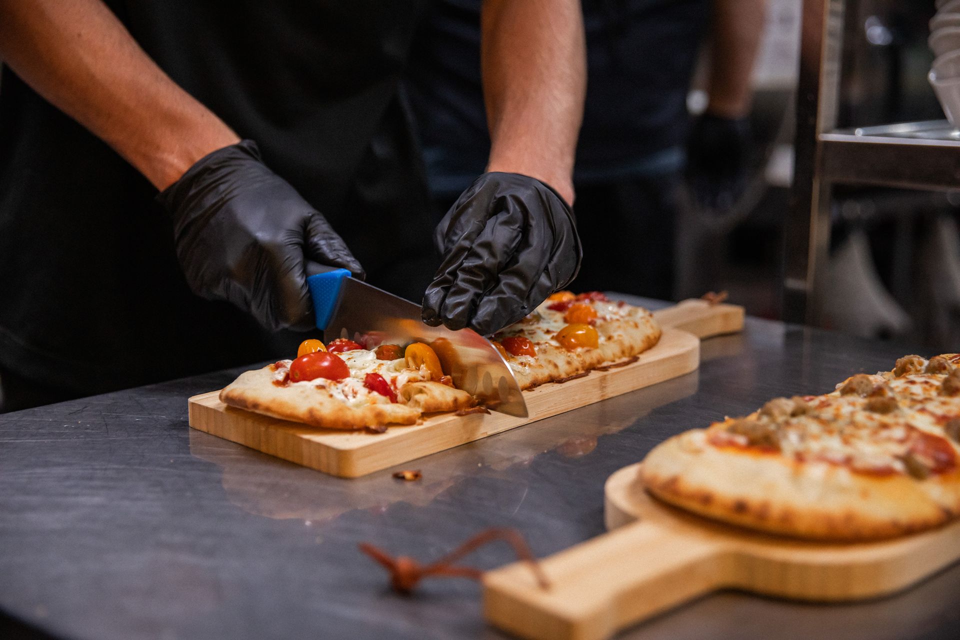 Chef slicing a pizza on a wooden board, wearing black gloves. Tomatoes and cheese visible. Kitchen setting.