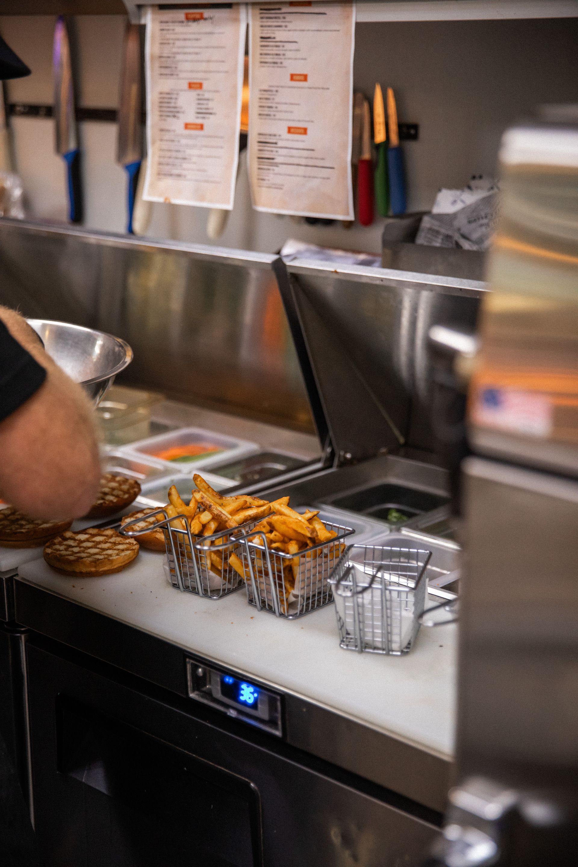 A person prepares food at a restaurant counter. Visible: fries, burgers, stainless steel surfaces, and menus.