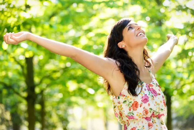 Woman with arms outstretched, smiling in a sunny green park, wearing a floral dress.