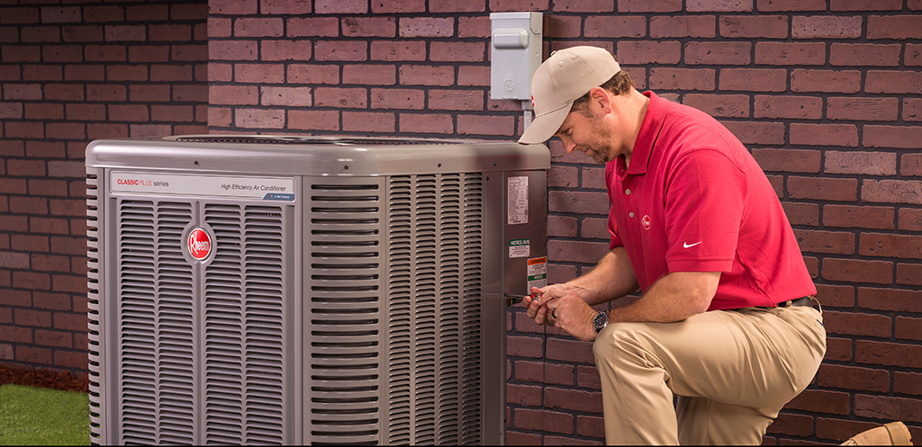 Person kneels to work on an air conditioner unit next to a brick wall.