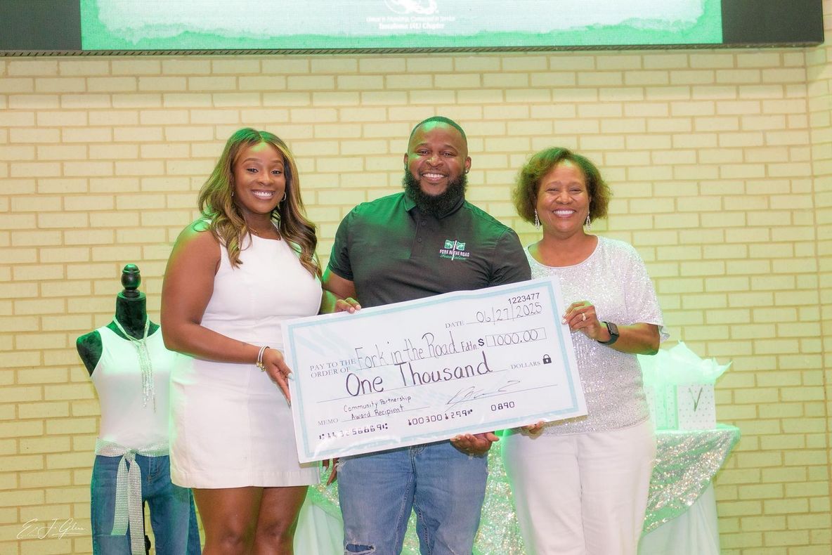 Three people hold a large check. Two women and a man smile, standing in a room with decorations and a display of clothing.