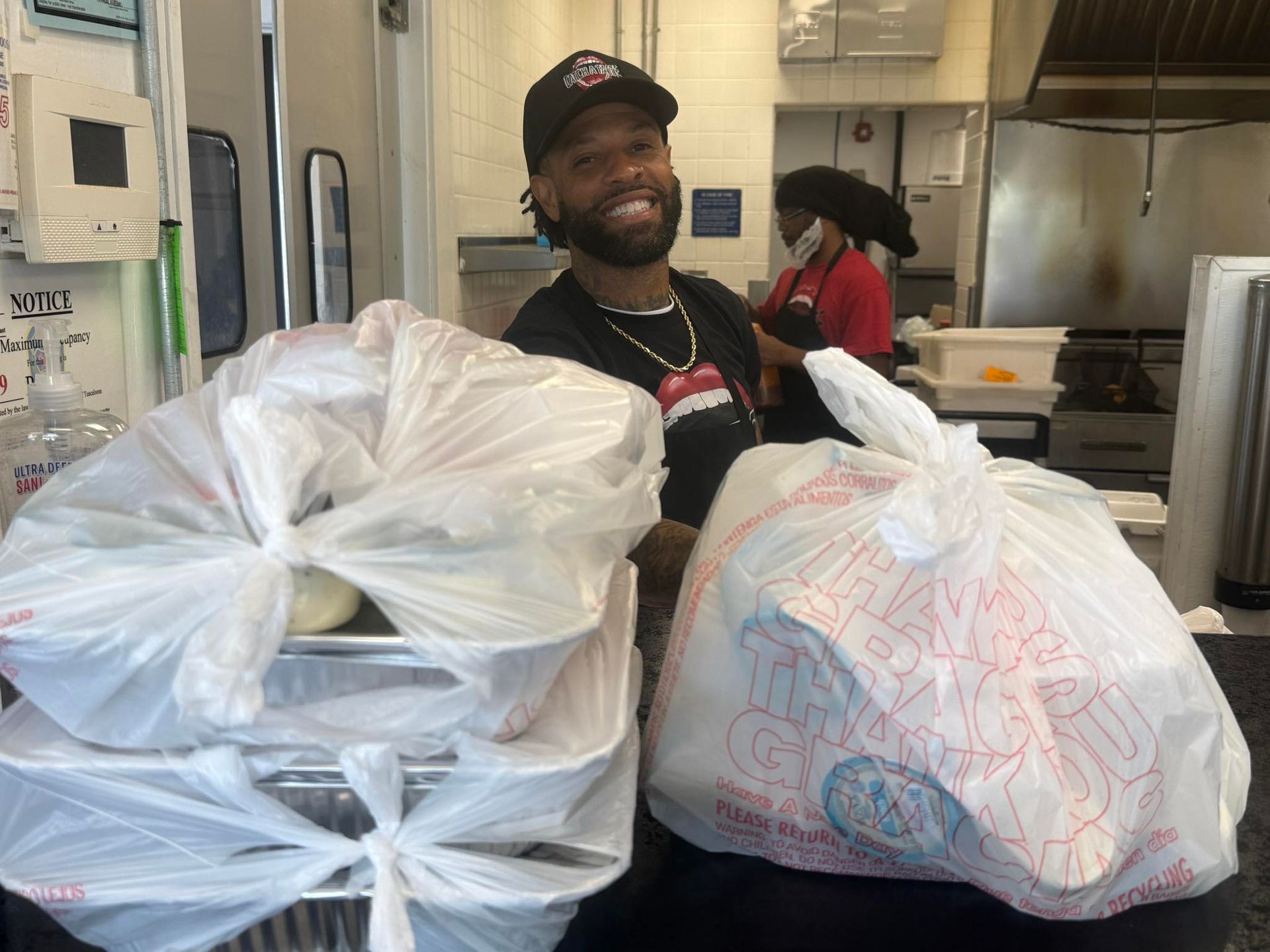 Man smiling with bags of food on counter in a restaurant kitchen. Another person in background.