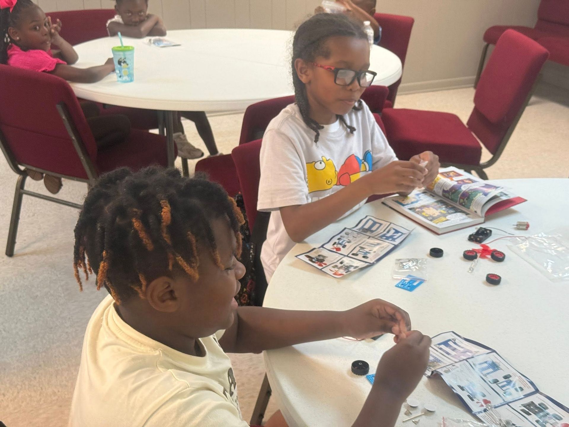 Children at white tables assembling toys from instructions. Red chairs, indoor setting.