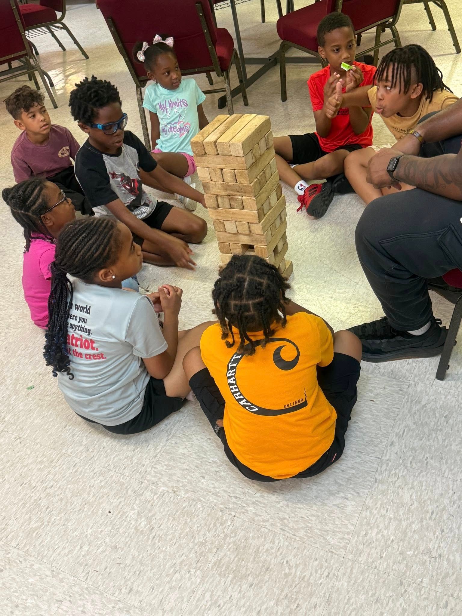 Children playing Jenga, sitting on the floor. A tall tower of blocks teeters; they watch intently. Indoors, room with chairs.