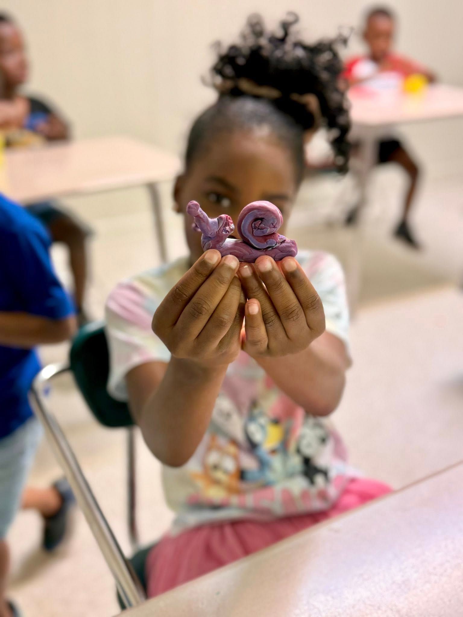 Girl holding up a purple clay snail creation in a classroom.