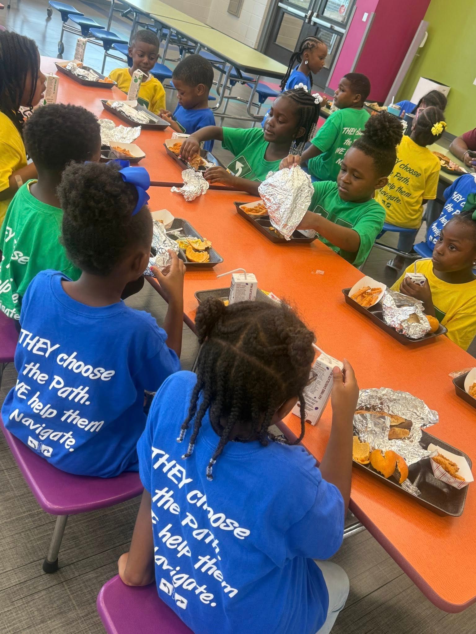 Children in green and blue shirts seated at a table, eating food wrapped in foil.