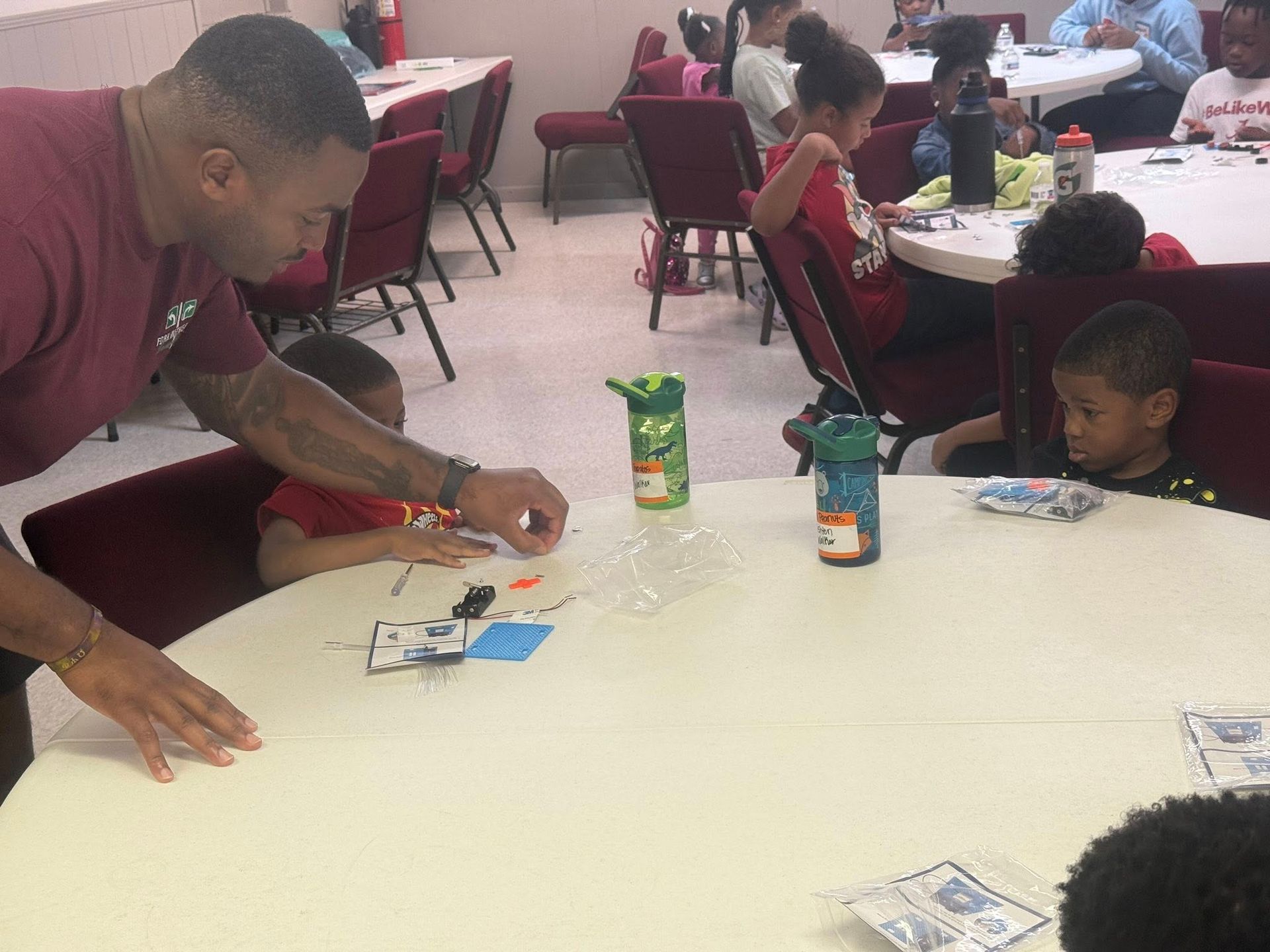 Adult assisting children at a table with a craft project, with others seated around the room.