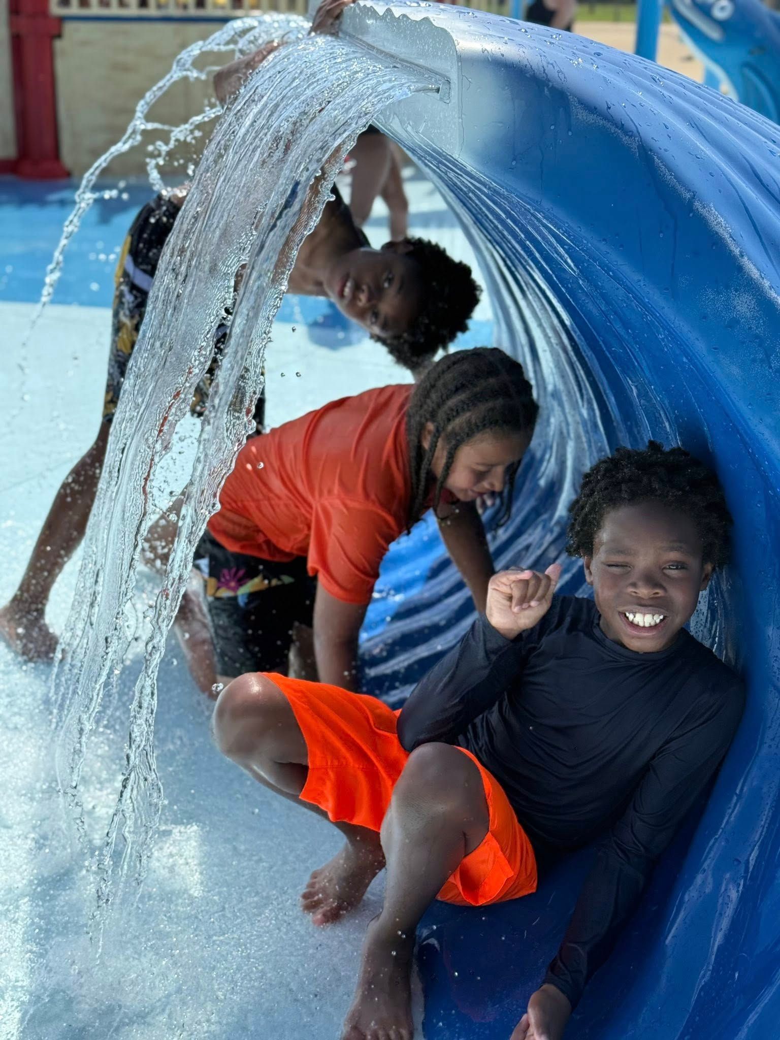 Children playing in a splash pad; water cascades over a blue tunnel. Smiling faces, orange and red swimsuits.