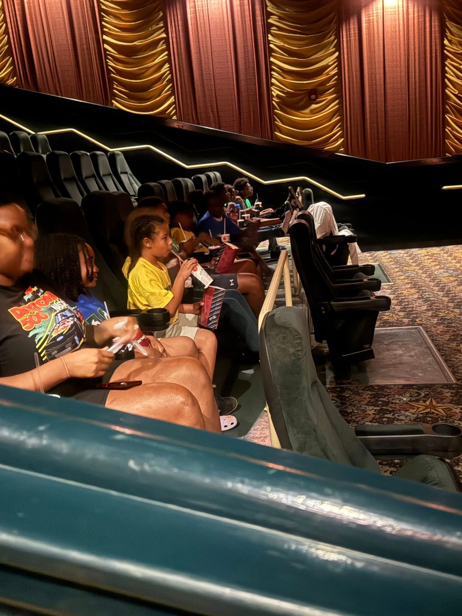 People seated in a movie theater, watching a film. Red curtains in the background.